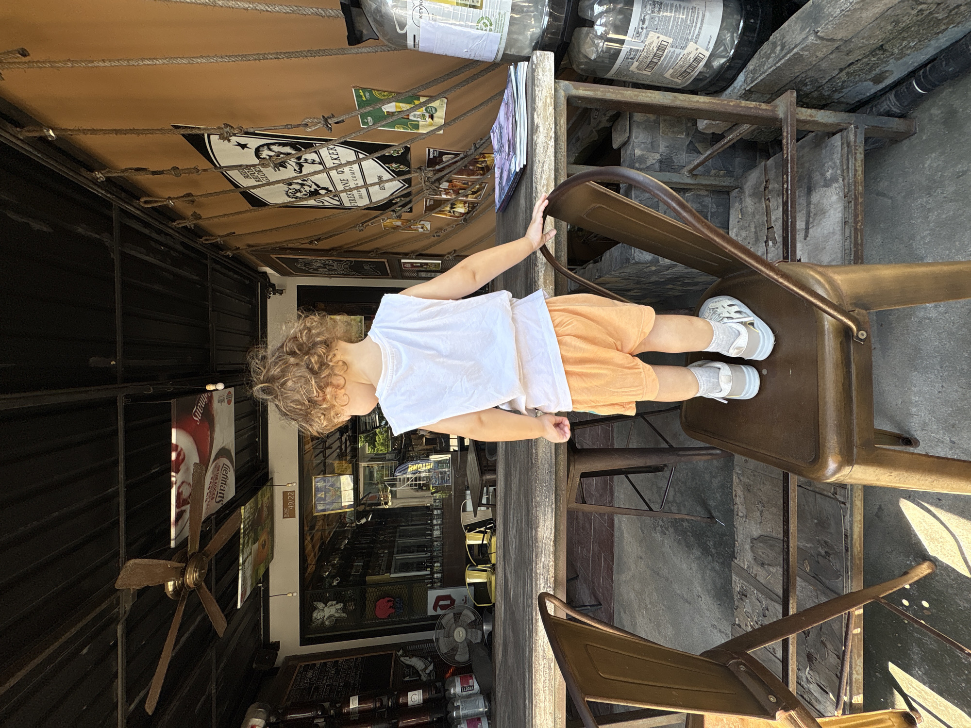 In the city of Choeng Thale, Thailand, a young child with curly blonde hair stands on a metal chair in front of a wooden table. The child is wearing a white sleeveless shirt and orange shorts. They are facing away from the camera, looking towards a building with an open-air facade that appears to be a bar or restaurant. The building has a corrugated metal roof and is decorated with various posters and signs. To the right of the child, there are large plastic containers. The scene is bathed in natural light, casting shadows on the ground.