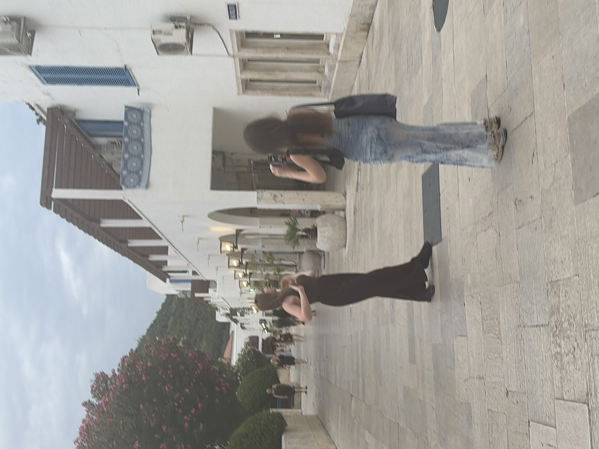In Budva, Montenegro, on a cobblestone street lined with white buildings adorned with wooden accents, a woman in a dark, flowing dress poses. She stands with her hands near her chest, her gaze directed away from the camera. Another woman, facing away, captures a photo with her phone while wearing a light blue denim-like dress and sneakers. She carries a dark tote bag over her shoulder. The street is illuminated by lampposts, and in the background, other pedestrians are visible. Lush green foliage and a hillside are present on the left side of the image. The sky above is overcast.
