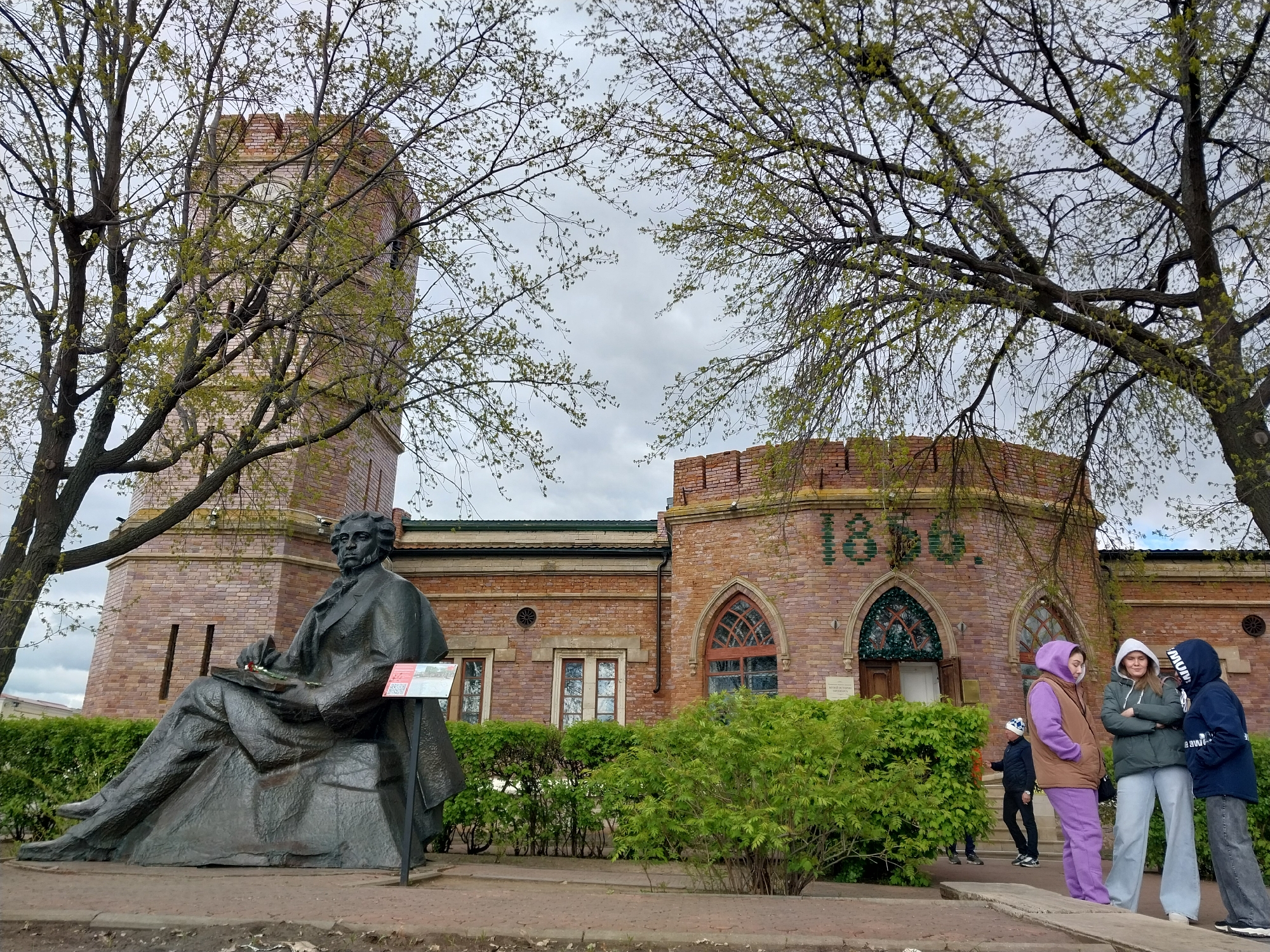 The image depicts the Museum of the History of Orenburg in Russia. A bronze statue of a seated man is in the foreground, positioned to the left of the museum building. The museum itself is constructed of red brick and features a tower on its left side, and an arched entrance displaying "1860" on the right. Three women stand to the right of the museum. One is wearing a purple sweatsuit with a brown vest, one is wearing a green jacket, and one is wearing a navy jacket and gray pants. A child is partially visible behind them. The weather appears overcast. Trees with budding leaves frame the scene on either side. A green hedge runs along the base of the museum and behind the statue.