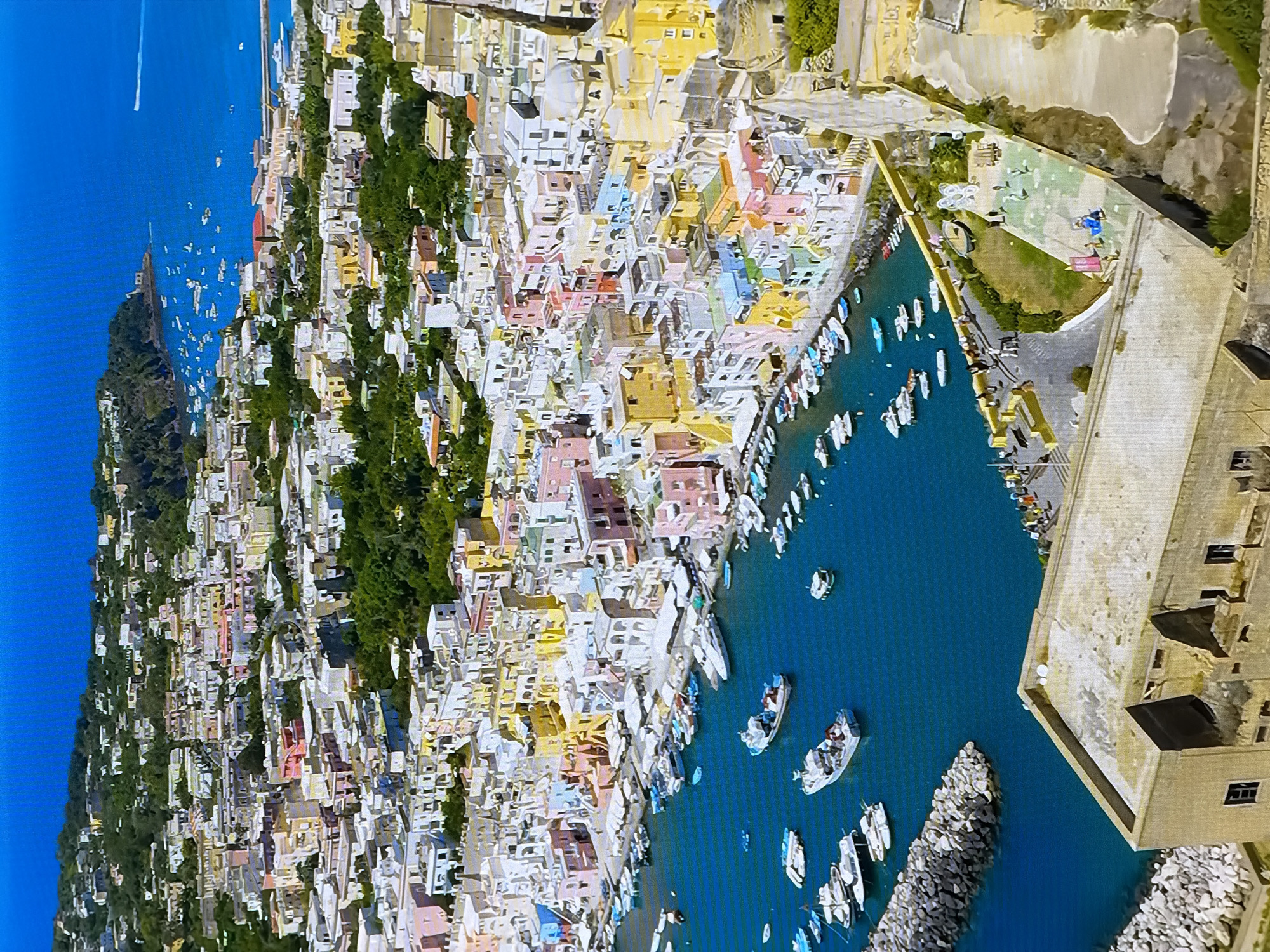This aerial photograph depicts a vibrant coastal town characterized by a densely packed collection of colorful buildings cascading down a hillside towards a harbor filled with numerous boats. The architecture is a mix of various sizes and shapes, with facades painted in shades of pink, yellow, blue, and white. Many buildings feature balconies and tiled roofs, contributing to the picturesque scene.
The harbor itself is a bustling hub with a variety of watercraft, including yachts, speedboats, and smaller fishing vessels, moored along the waterfront. The water is a deep, clear blue. A breakwater, constructed from large rocks, extends into the harbor, providing a protective barrier.
Beyond the immediate waterfront, the town extends further inland, with more buildings interspersed with lush green vegetation and trees. The hilly terrain suggests a natural landscape integrated with the urban development. The sky is a bright, cloudless blue, indicating a clear, sunny day.
The lower portion of the image focuses on a large, aged structure, possibly a historical building or fortification, with a weathered facade and visible interior spaces. A pathway leads from this structure towards the waterfront, where several individuals are seen walking and interacting.
The scene is located in the city of Chirchik, Uzbekistan.