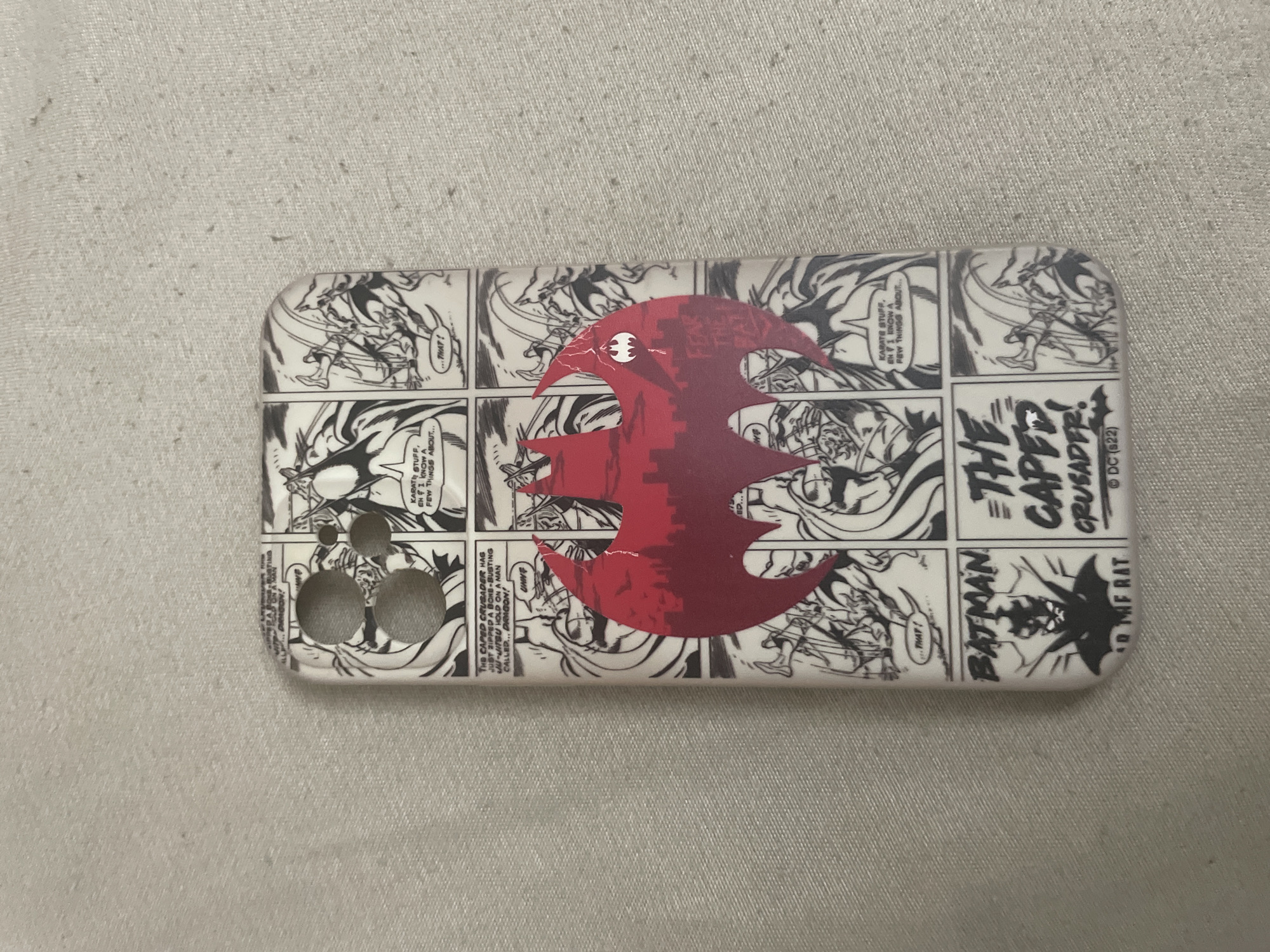 A white smartphone case featuring a black and white comic book panel design is centrally positioned on a light gray, textured fabric surface. The case has three circular cutouts for a camera module in the upper left section. A prominent red Batman symbol with a silhouette of a city skyline at its base is overlaid on the central panels. Within the red symbol, a smaller white Batman emblem is visible.
The comic panels depict various action scenes with characters in a dynamic, hand-drawn style, primarily in black and white. Visible text snippets from speech bubbles and captions include "KARATE STUFF, EH? I KNOW A FEW THINGS ABOUT...", "...THAT!", "THE CAPED CRUSADER HAS JUST ZIPPED A BONE-BUSTING JU-JITSU HOLD ON A MAN CALLED... DRAGON!", "UHWE", "FEAR THE BAT", "BATMAN THE CAPED CRUSADER!", "AD THE RAT", and "© DC (S22)". The fabric background exhibits a consistent, fine-grained weave with subtle variations in shade.