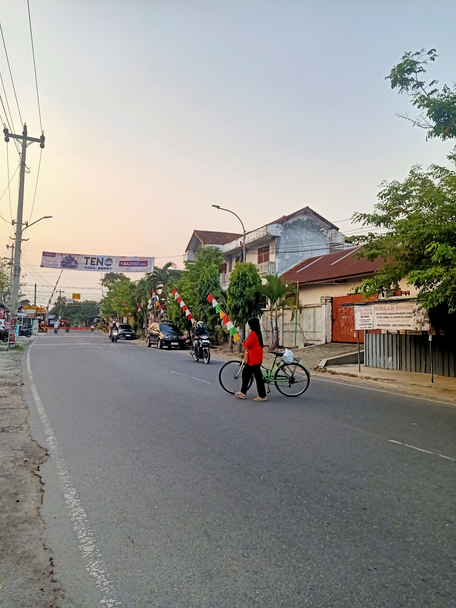 The image depicts a street scene in Klaten, Indonesia, at dusk. A woman in a red shirt and black pants walks her bicycle across a paved road. The road has a white dividing line. In the background, buildings with red-tiled roofs are visible, along with trees and overhead power lines. A banner advertising "TEN mii COOL BERRY" is strung across the road. Several motorcycles and cars are also present on the road, and a railway crossing barrier is visible on the left side of the image. The sky is a soft gradient of orange and light blue, indicating the setting sun. A sign on a building to the right reads "BERKAH SNACK" and lists various food items.