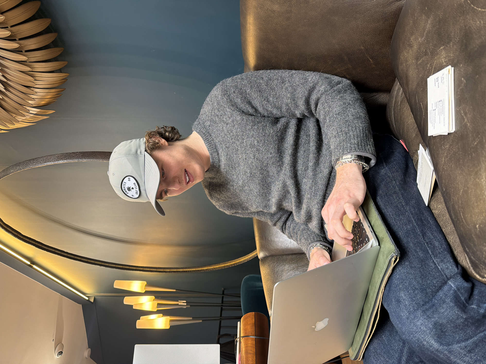 A young adult male, with curly brown hair partially visible, is seated on a dark brown leather couch in a modern lounge or lobby area in Paris, France. He wears a grey baseball cap with an "EMIRATES 88 GOLF CLUB" patch, a grey knit sweater, and dark blue jeans. His left wrist displays a silver chain-link watch. He is actively typing on an open silver Apple MacBook laptop, which rests on an olive green zippered laptop sleeve on his lap. To his right, on the couch beside his leg, a small stack of white business cards or papers is visible. The background features a teal-colored wall adorned with a large, dark circular mirror or art piece and a golden, leaf-shaped sculptural decoration. Warm ambient lighting emanates from a modern four-stemmed floor lamp with conical shades and a linear LED strip along the ceiling. A white dome-shaped security camera is mounted on the ceiling in the upper left. A small wooden table and a partial view of teal upholstery are visible in the left background.