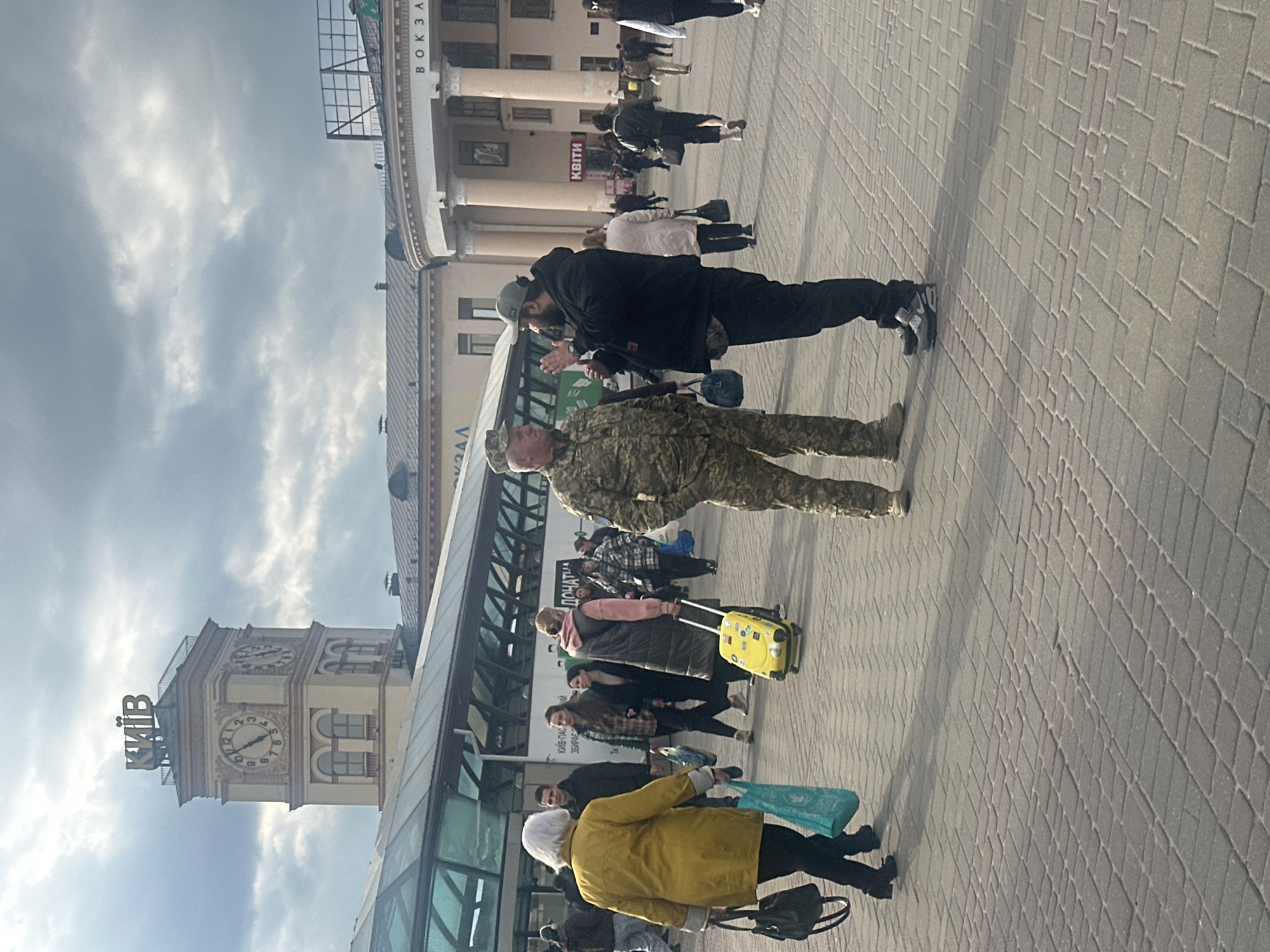 The image captures an outdoor scene in Kyiv, Ukraine, under a partly cloudy sky, likely during the daytime. The perspective is that of someone standing on a paved area, looking towards several buildings.
In the foreground, a person wearing a full military camouflage uniform, consistent with Ukrainian military attire, stands facing a civilian man. This civilian wears a black hooded jacket, dark pants, and a grey baseball cap, and has a beard. His hands are slightly raised, as if in conversation or greeting.
Behind these two figures, numerous other civilians are visible, walking across the paved ground, which is made of light grey pavers. Some individuals are carrying luggage; notably, a person in the mid-ground pulls a bright yellow wheeled suitcase. Another woman in a yellow coat carries a light blue tote bag and a dark handbag. The overall activity suggests people are moving through a public space, possibly arriving at or departing from a transportation hub.
The background features prominent architectural structures. To the left, a tall, beige clock tower stands against the sky, with the word "КИЇВ" (Kyiv) written in large letters at its top. The clock faces are visible, with the time appearing to be around 8:50 or 9:50. To the right, a grand building with classical columns can be seen. Above these columns, the word "ВОКЗАЛ" (Vokzal), meaning "Station," is clearly visible, identifying the building as a train or bus station. Another sign on a building further to the right reads "КВІТИ" (Kvitky), meaning "Flowers."
The scene is bustling, reflecting everyday life and travel in the city, with the presence of military personnel adding a contextual layer related to the current situation in Ukraine.