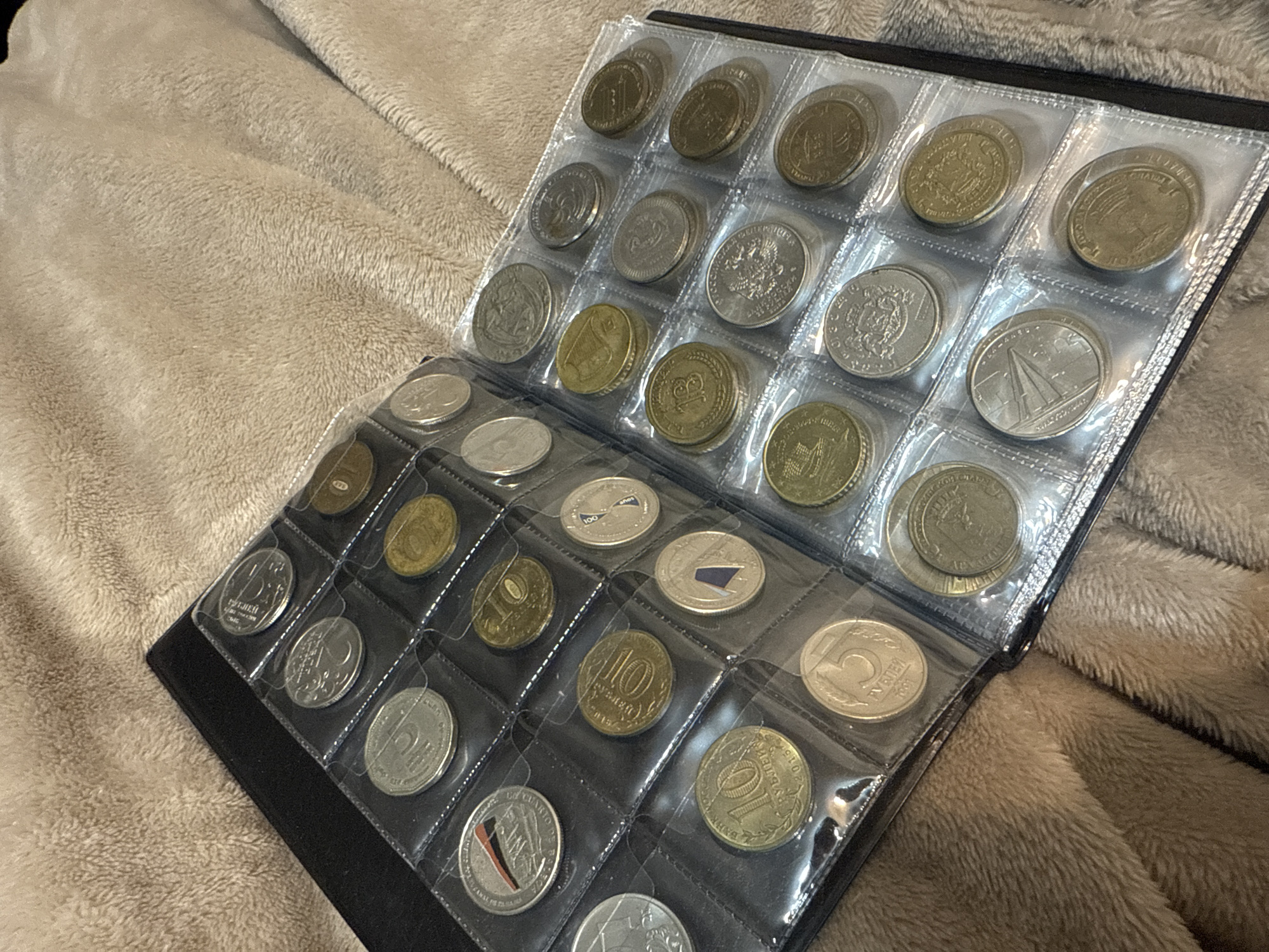 The image shows a black coin collecting album open on a beige, plush surface. The album contains numerous coins encased in clear plastic sleeves, arranged in a grid. The coins are of varying colors (silver, gold, bronze) and designs. The album appears to be lying on a soft, textured blanket or throw. The overall composition is close-up and angled, providing a detailed view of the coins and album within the context of the surrounding plush material. It is not possible to determine the location in the city of Mytishchi, Russia, from the image provided.