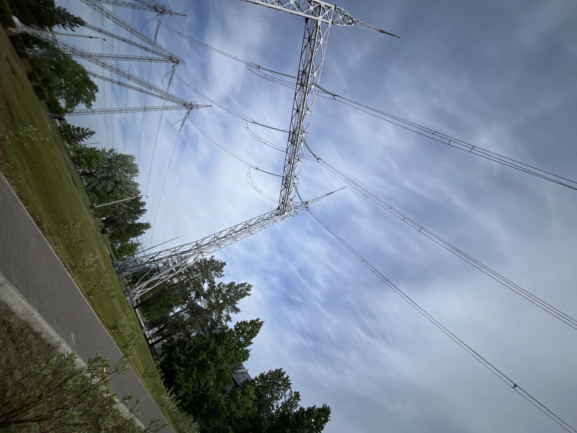 The image shows a power line structure in Chaikovsky, Russia. Several lattice-structured steel transmission towers stand in a grassy area bordered by trees and a paved walkway. Power lines stretch across the sky, which is filled with cirrus clouds. The viewpoint is low, looking upwards at an angle, emphasizing the height and scale of the power infrastructure.