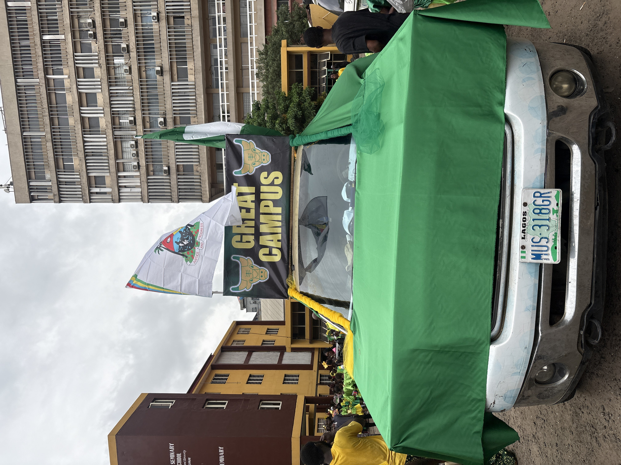 The image shows a vehicle in Lagos, Nigeria. The vehicle has the license plate "MUS-318GR" and is draped with green fabric, with the words "GREAT CAMPUS" displayed on a banner. People are gathered around the vehicle, some wearing green and yellow clothing. Buildings are visible in the background, including one with multiple windows and air conditioning units. Another building in the background has a sign that reads "SEMINARY SCHOOL of and Sincerity ALUMNI". There are multiple flags; one is the flag of Nigeria. The other flag also contains an emblem of a bull.