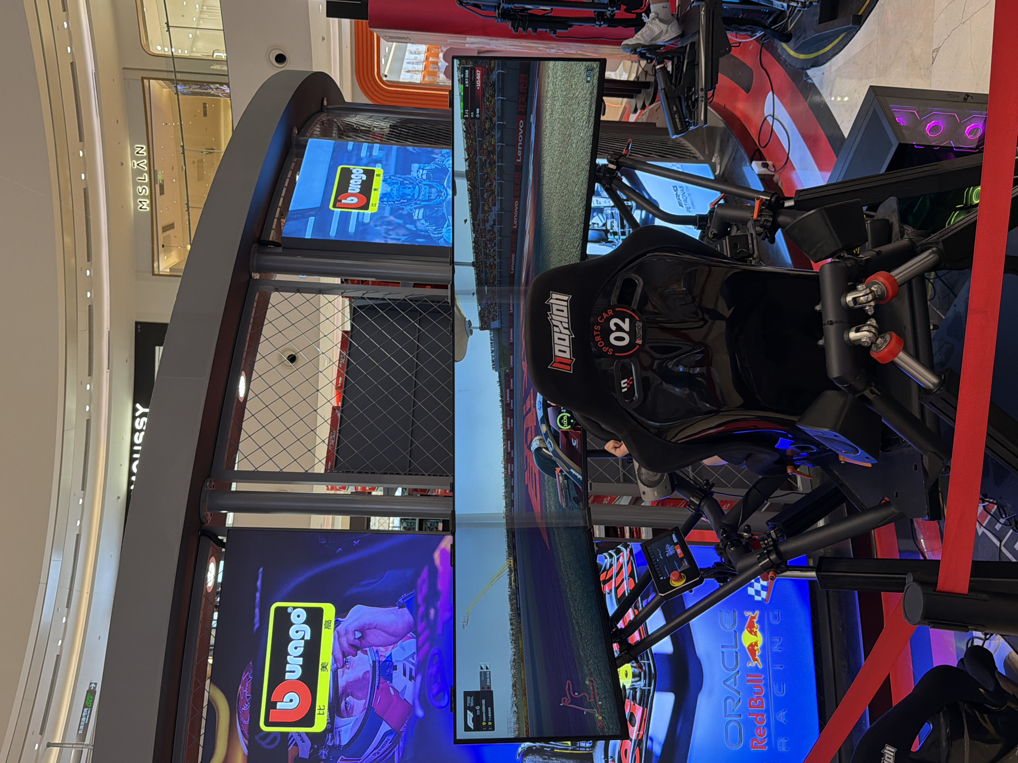 The image captures a detailed view of a professional-grade racing simulator setup located within a modern, brightly lit shopping mall in Yuzhong District, China.
At the center, a black racing simulator rig features a contoured racing seat with "NOOKXION" and "SPORTS CAR 02" branding. A person is seated in the simulator, with their right hand visible on a control stick, operating the system. The rig is equipped with a triple-monitor display setup, presenting a wide-angle view of a Formula 1 racing game track, complete with virtual trackside banners featuring "Lenovo" and "联想" (Lenovo in Chinese) branding, spectator stands, and in-game telemetry like lap times and positions. The simulator's control panel, featuring buttons and a small screen, is mounted to the left of the driver. A high-end PC tower with purple LED lighting powers the system, visible on the right. Red velvet ropes delineate the public access area around the simulator. The flooring beneath the setup is designed with red and black stripes, resembling a racetrack.
Above the simulator, a series of large vertical LED screens display the "Bburago" logo (a red "b" in a yellow rectangle) alongside Chinese characters "比美高" (Bǐměigāo). One screen prominently features an image of a racing driver wearing a helmet with "Verstappen" visible, while another screen on the far left shows the "Oracle Red Bull Racing" logo.
The surrounding environment is a multi-story shopping mall characterized by a curved, white ceiling with numerous recessed lights. On an upper level, storefronts for "MOUSSY" and "MSLAN" are visible. A directional sign with Chinese characters and an arrow is mounted on a wall above the Bburago screens on the left. Another person's foot, clad in a white sneaker, is partially visible on the far right of the image, near additional equipment.