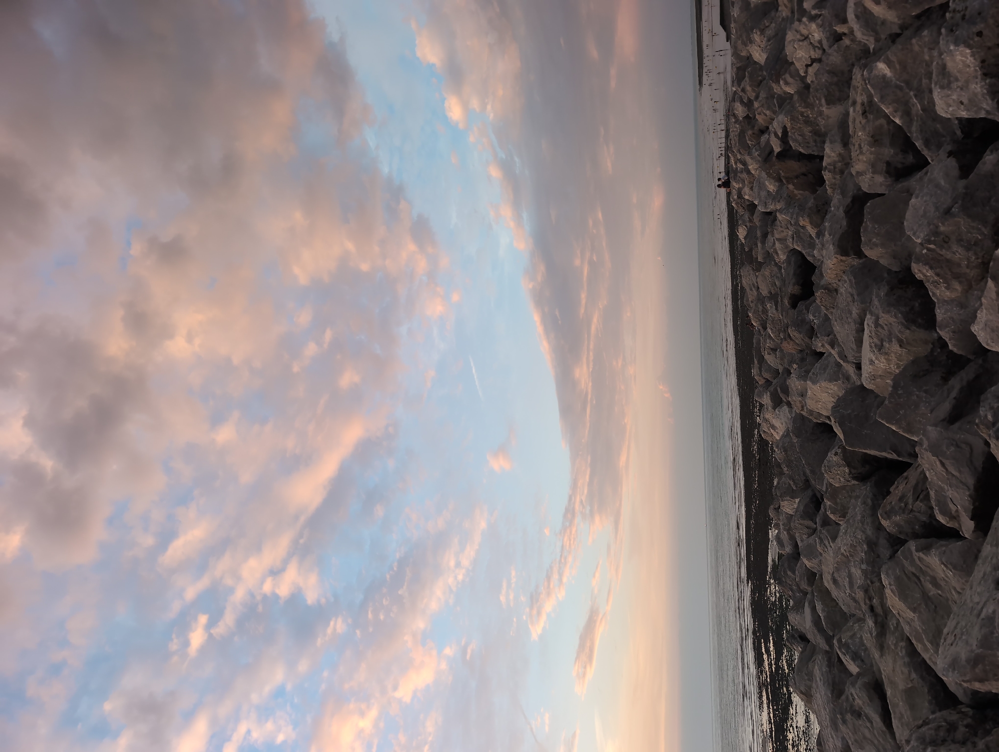 The image depicts a view of the rocky shoreline in Le Tréport, France, with the English Channel in the background and a pastel-colored sky overhead. The foreground is dominated by a dense arrangement of large, grey rocks, forming a breakwater. The sea stretches to the horizon, appearing calm and reflecting the soft hues of the sky, which is filled with streaks of pink, orange, and blue clouds. A faint line of people and structures can be seen in the distance along the coast, suggesting the presence of a town or promenade. The overall impression is one of a serene coastal landscape at dusk.