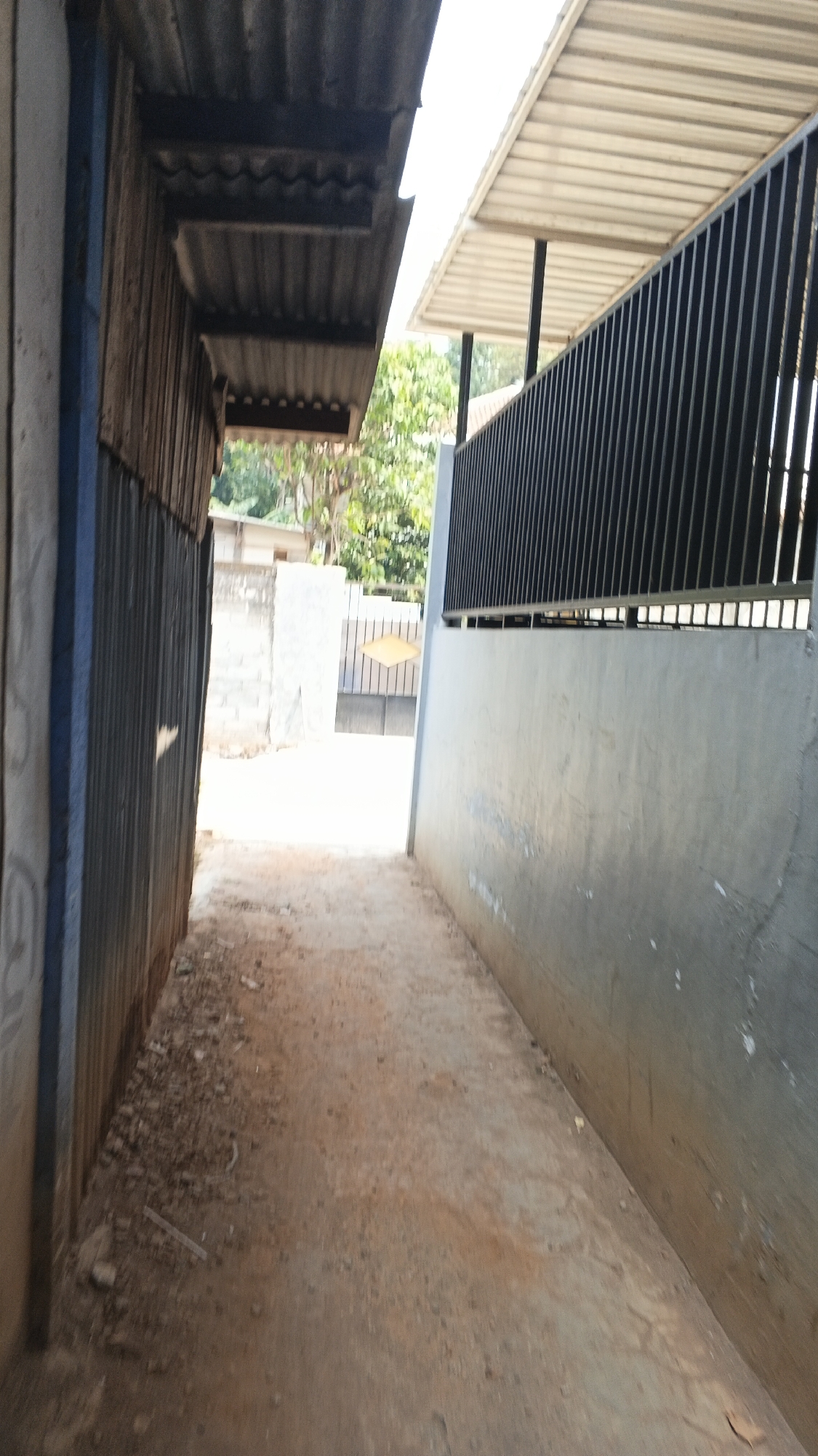 The image shows a narrow outdoor alleyway between two buildings. The building on the left has a corrugated metal wall and a corrugated metal roof. The building on the right has a concrete wall and a black metal fence with a corrugated metal roof. The ground in the alleyway is covered with dirt and small rocks. In the background, there is a gate with a diamond pattern and some trees and foliage. The lighting suggests it is daytime. There are no people or animals visible in the image.