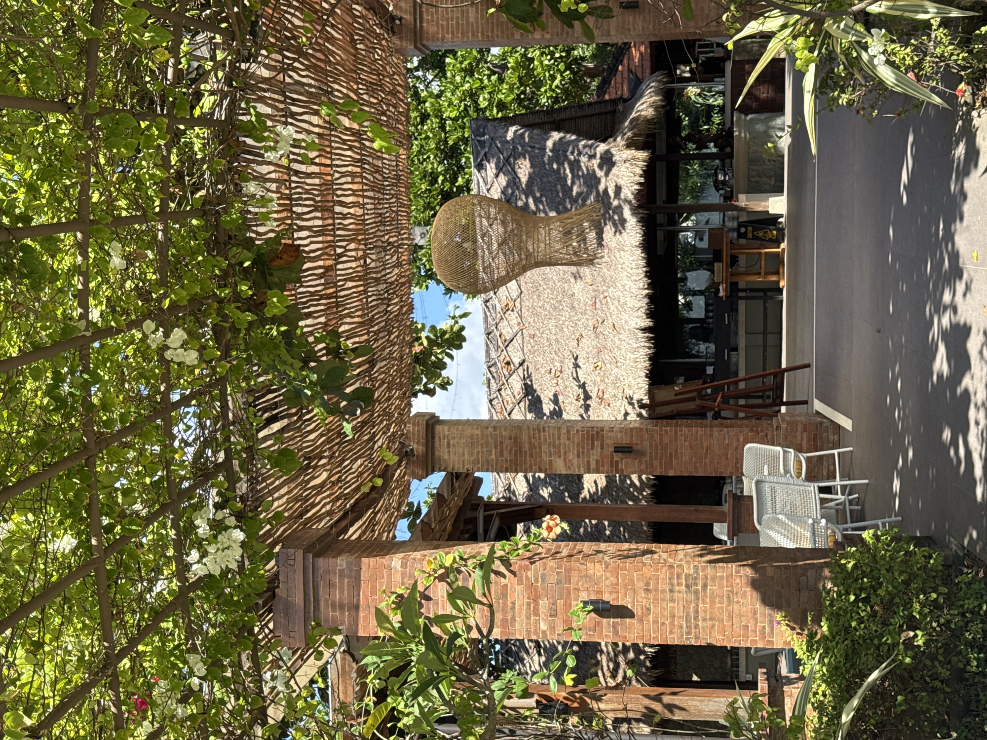 The image, taken in Kuta Selatan, Indonesia, shows a partially covered outdoor seating area. The roofing consists of a thatched material over brick pillars. Plants with green foliage are seen in the foreground. Adjacent to the seating area is a paved surface. The image also reveals an exterior building with tables and chairs visible. An unusual woven light fixture hangs from the thatched roof.