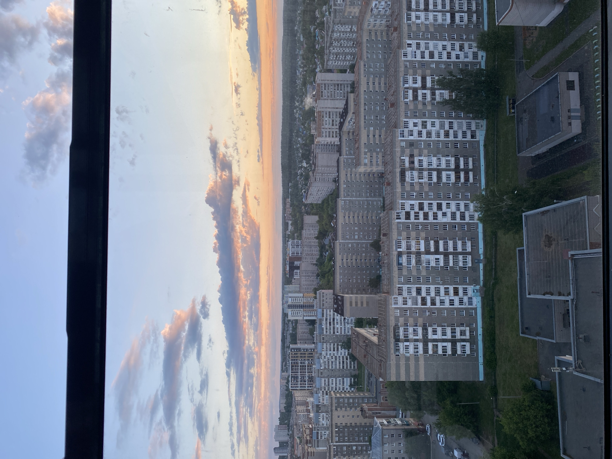 This image presents a panoramic view of a city at either sunrise or sunset, captured from a high vantage point, likely through a window. A dark, vertical window frame runs along the left side of the composition.
The cityscape in the midground features numerous multi-story residential buildings, primarily characterized by grey facades and repetitive patterns of white-framed windows and balconies, typical of Soviet-era or post-Soviet architecture. Green spaces, including strips of grass and scattered trees, are visible between the buildings, adding natural elements to the urban environment. Several smaller, darker buildings with flat roofs, possibly garages or commercial structures, are also interspersed. A road or pathway with a few parked cars can be seen weaving through the urban fabric.
In the background, the city extends into the distance, meeting a distinct horizon line marked by a dense band of trees or forest. The sky is a prominent feature, displaying the warm, vibrant colors of a dawn or dusk. Scattered clouds near the horizon are illuminated with shades of orange, pink, and purple, gradually transitioning into a lighter blue sky overhead. The light bathes the entire scene in a soft, golden glow.
No specific human activity or visible text is discernible in the image. The architectural style and urban layout are consistent with the provided location context of Izhevsk, Russia. The overall impression is one of a tranquil urban landscape observed during a beautiful atmospheric event.
