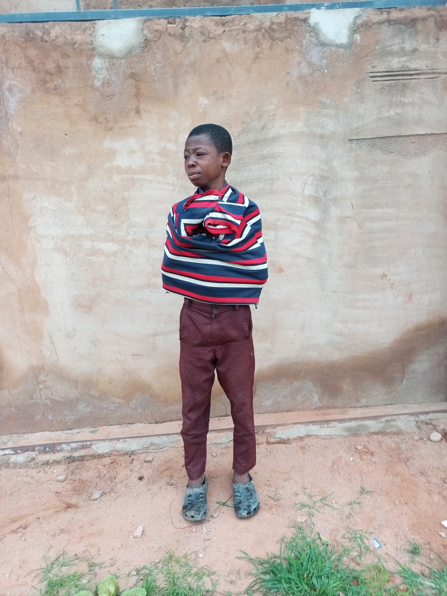 A young boy stands with his arms crossed over his chest, wrapped in a striped garment. He is wearing maroon pants and dark, worn-out clogs. The boy faces away from the camera, looking to his left with a pensive expression. He stands on sandy ground in front of a textured, weathered wall. The wall shows signs of age with cracks and discoloration. A blue railing runs horizontally along the top of the wall. In the foreground, patches of green grass are visible, with a few small green objects on the ground. The scene appears to be outdoors in a simple, unadorned setting.