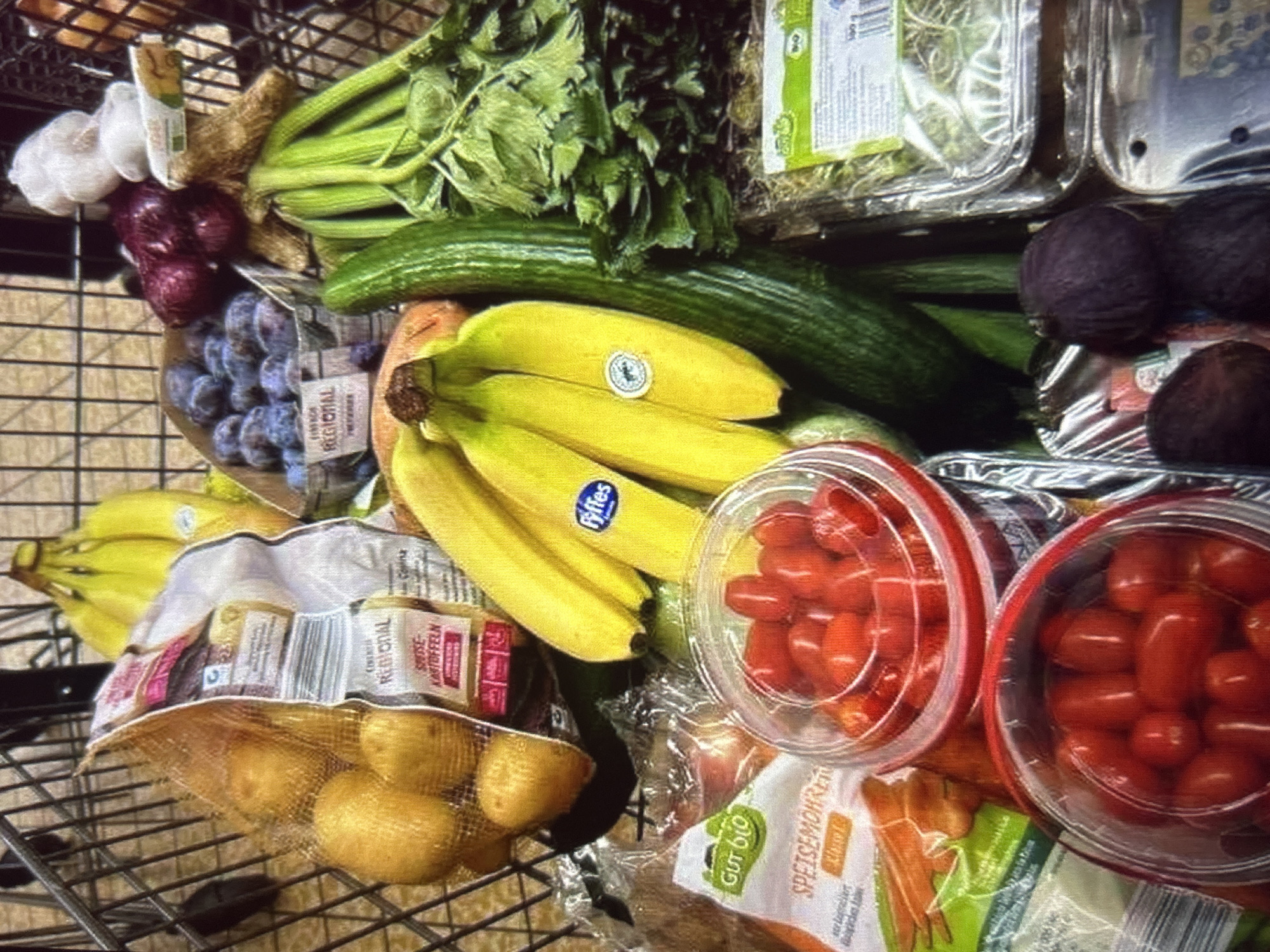 The image shows a close-up, overhead view of a shopping cart filled with groceries. The items include potatoes in a mesh bag, a bunch of bananas with a Fyffes sticker, a container of cherry tomatoes, a bag of carrots, a red onion, garlic, plums in a cardboard container, celery, and a cucumber. The background is out of focus, but appears to be a grocery store aisle. There are no people or specific landmarks visible in the image, making it impossible to determine the location as Knezha, Bulgaria.
