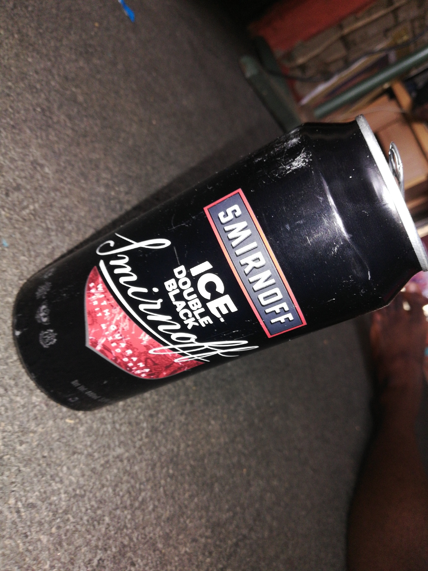 A slightly damaged black can of Smirnoff Ice Double Black lies on a textured gray surface. The can displays the "Smirnoff" logo in white cursive script and a red patterned area. The can is slightly tilted, with the pull tab visible. A small portion of a dark-skinned human foot is visible in the lower right corner of the frame. The background suggests an indoor setting. The city is Unknown, Unknown.