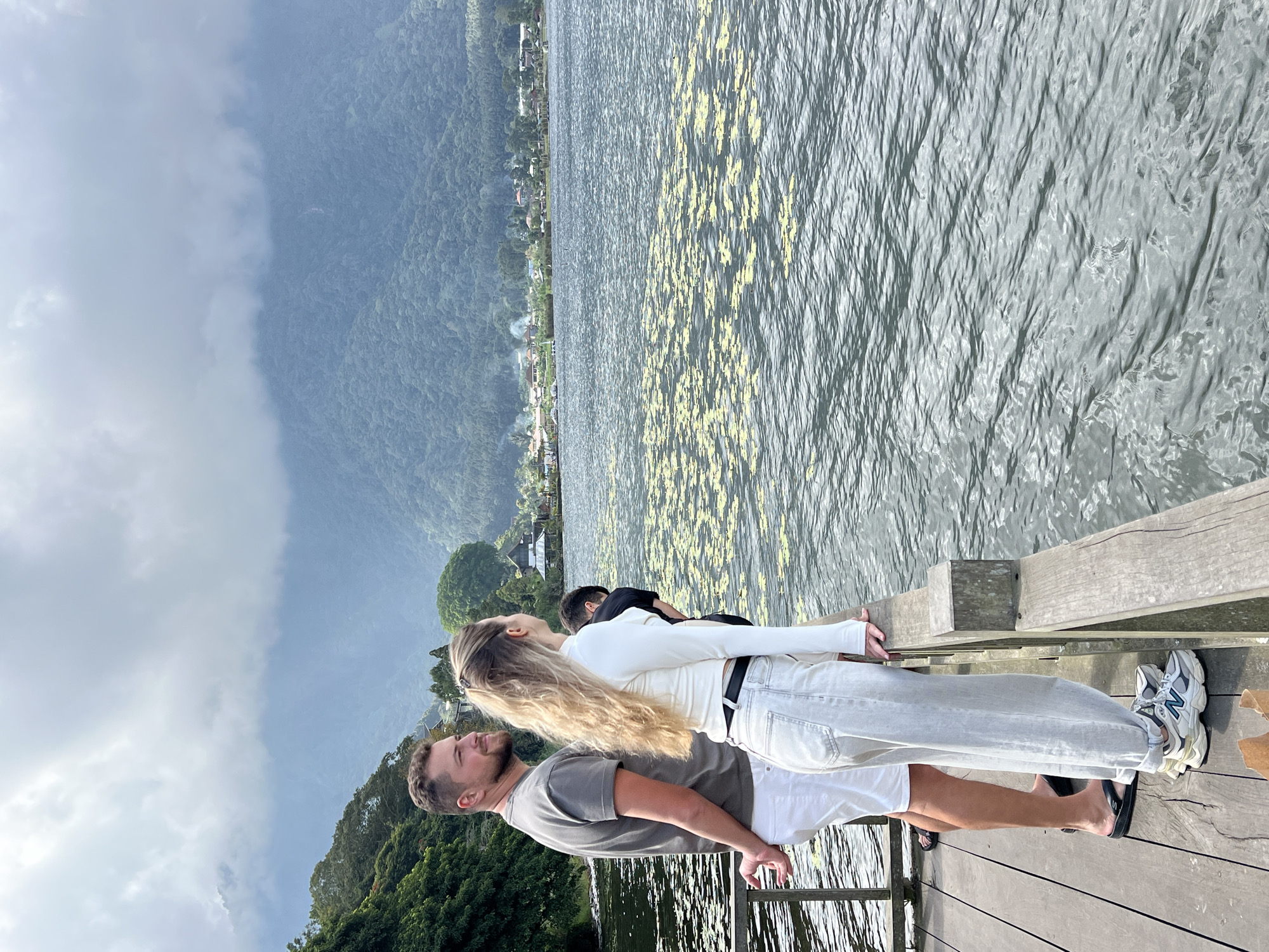 A man and a woman stand on a wooden dock, looking out at a lake. The man is wearing a gray t-shirt and white shorts, with black flip-flops. The woman is wearing a white long-sleeved shirt and light-colored pants, with athletic shoes. Behind them, a third person, partially obscured, stands on the dock. The lake is a dark gray color with patches of yellow lily pads floating on the surface. In the background, a lush green mountainside is visible, with a small village nestled at its base. The sky is overcast with heavy gray clouds. The image appears to have been taken in Baturiti, Indonesia, specifically near Lake Batur, known for its volcanic crater lake and surrounding mountain scenery.