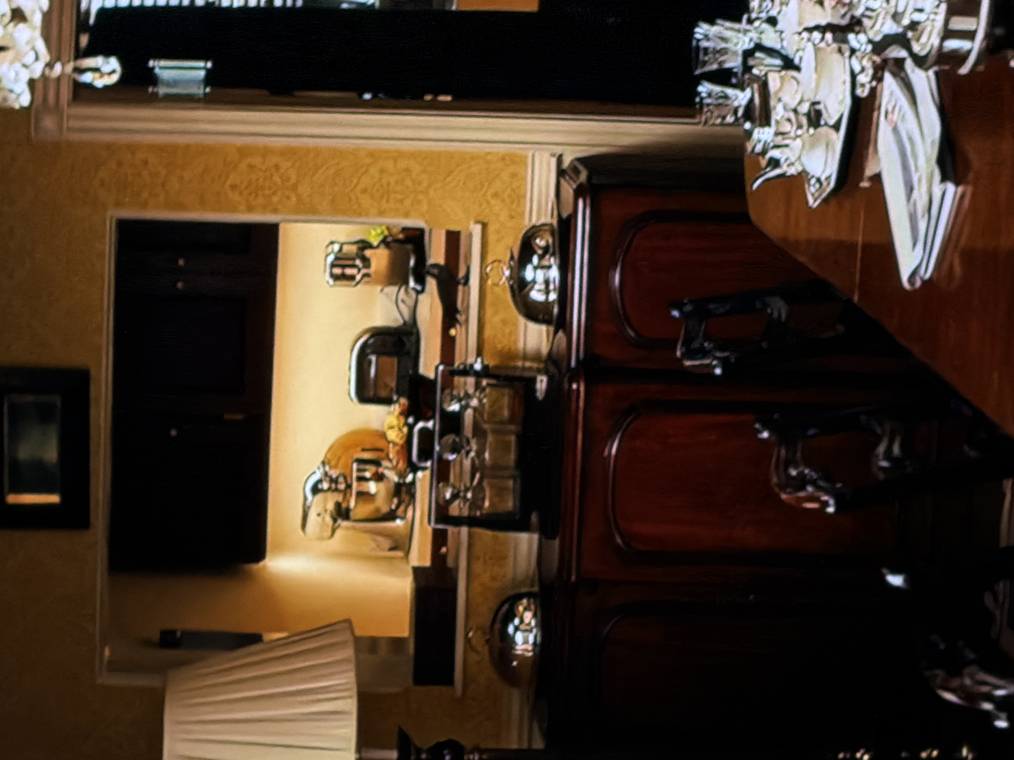 This indoor scene depicts a formal dining or buffet area, likely in a luxurious home, restaurant, or hotel in Sofiivska Borshchahivka, Ukraine. The image is taken from one room looking into an adjacent one, both furnished with elegant decor.
In the foreground to the right, a dark wooden dining table is visible, meticulously set with multiple white plates, glasses, and cutlery, suggesting preparation for a meal or an ongoing event. Adjacent to the table, a dark, ornate wooden cabinet or sideboard holds several silver-colored serving dishes with domed lids, typically used as chafing dishes to keep food warm in a buffet setup.
Through a doorway or archway, an adjacent room is visible. This room also features similar shiny, metallic serving dishes on a surface, further emphasizing a buffet-style arrangement. To the far left in this adjacent room, a dark rectangular object, possibly a framed picture or a mirror, hangs on the wall. In the immediate foreground to the left, a white pleated lampshade from a table lamp adds to the ambient lighting.
The walls throughout the visible spaces are a textured golden or beige color, adorned with a subtle patterned wallpaper, contributing to the opulent atmosphere. The lighting is dim, suggesting an evening setting or controlled indoor illumination. No people are present, and there is no visible text. The overall ambiance is one of quiet elegance and preparedness for a formal gathering.