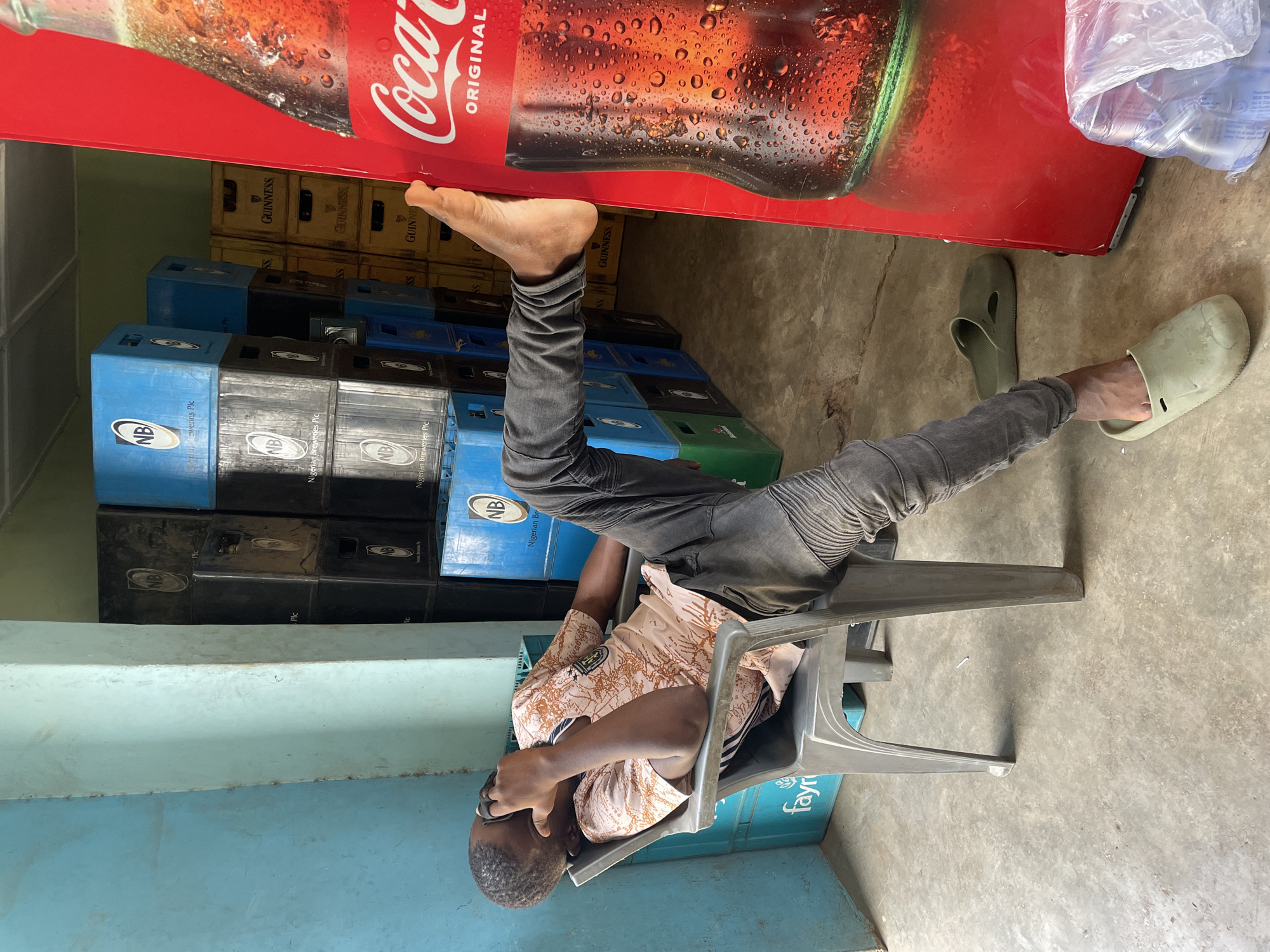 The image, rotated 90 degrees clockwise, captures a man resting on a grey plastic chair within what appears to be a small shop or beverage storage area in Jos, Nigeria. The man, with dark skin and short dark hair, is wearing a patterned light brown and black short-sleeved shirt and dark grey jeans. He is slumped in the chair with his head bowed, indicating he is either asleep or resting deeply. One bare foot is propped up against a red Coca-Cola refrigerator, while the other foot, wearing a light green slide sandal, is on the concrete floor beside its match.
Behind the man and the refrigerator, numerous beverage crates are stacked. Many blue crates prominently feature the "NB" logo and "Nigerian Breweries Plc" text. Yellow and black crates display "GUINNESS" and "SS". A partial text "fayri" is also visible on a crate near the chair. On top of the red Coca-Cola refrigerator, which showcases a large image of a cold, condensation-covered Coca-Cola bottle with "Coca-Cola" and "ORIGINAL" branding, are clear plastic bags, possibly containing ice or water sachets. The background includes a light blue painted wall or ledge, and the floor is concrete. The scene portrays a moment of quiet repose in a commercial setting.