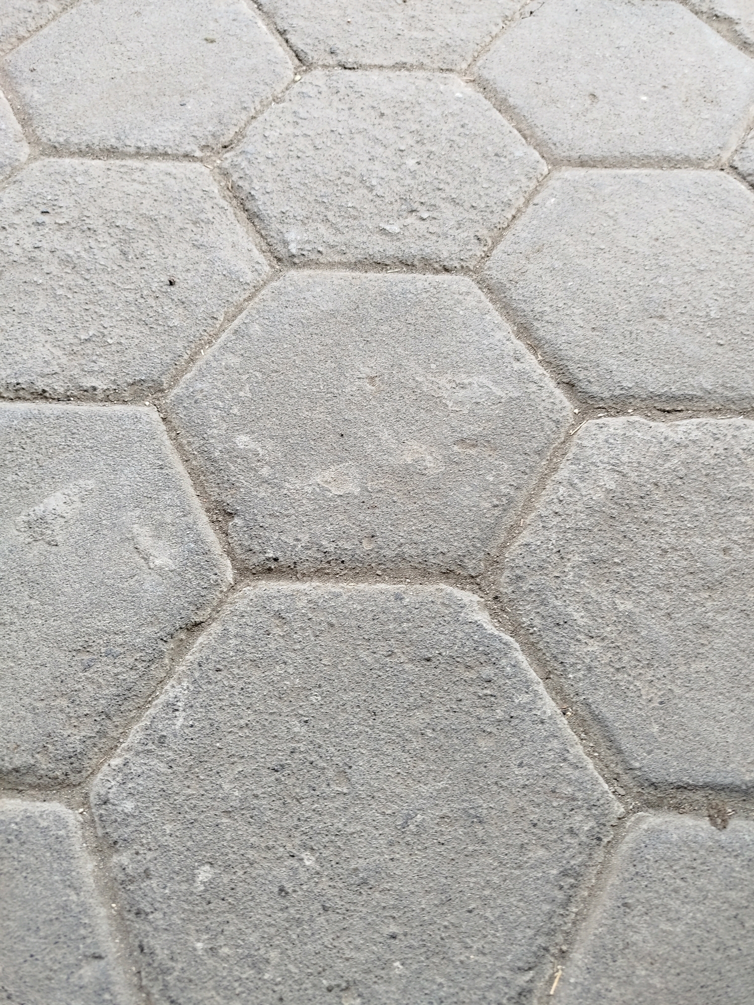 The image shows a close-up, overhead view of hexagonal paving stones, commonly known as "paving blocks" or "conblok". These are gray, interlocking concrete units laid out in a repeating hexagonal pattern. The texture of the stones is rough and gritty, with small dark aggregate visible on the surface. The grout lines between the stones are visible, filled with sand or fine gravel, and appear slightly worn. There is some minor discoloration and debris on the surface of the paving blocks, suggesting they are part of an outdoor walkway or paved area. The scene is consistent with paving found in public spaces in Karangwuni, Indonesia, such as sidewalks or pedestrian areas. No people or specific landmarks are visible.