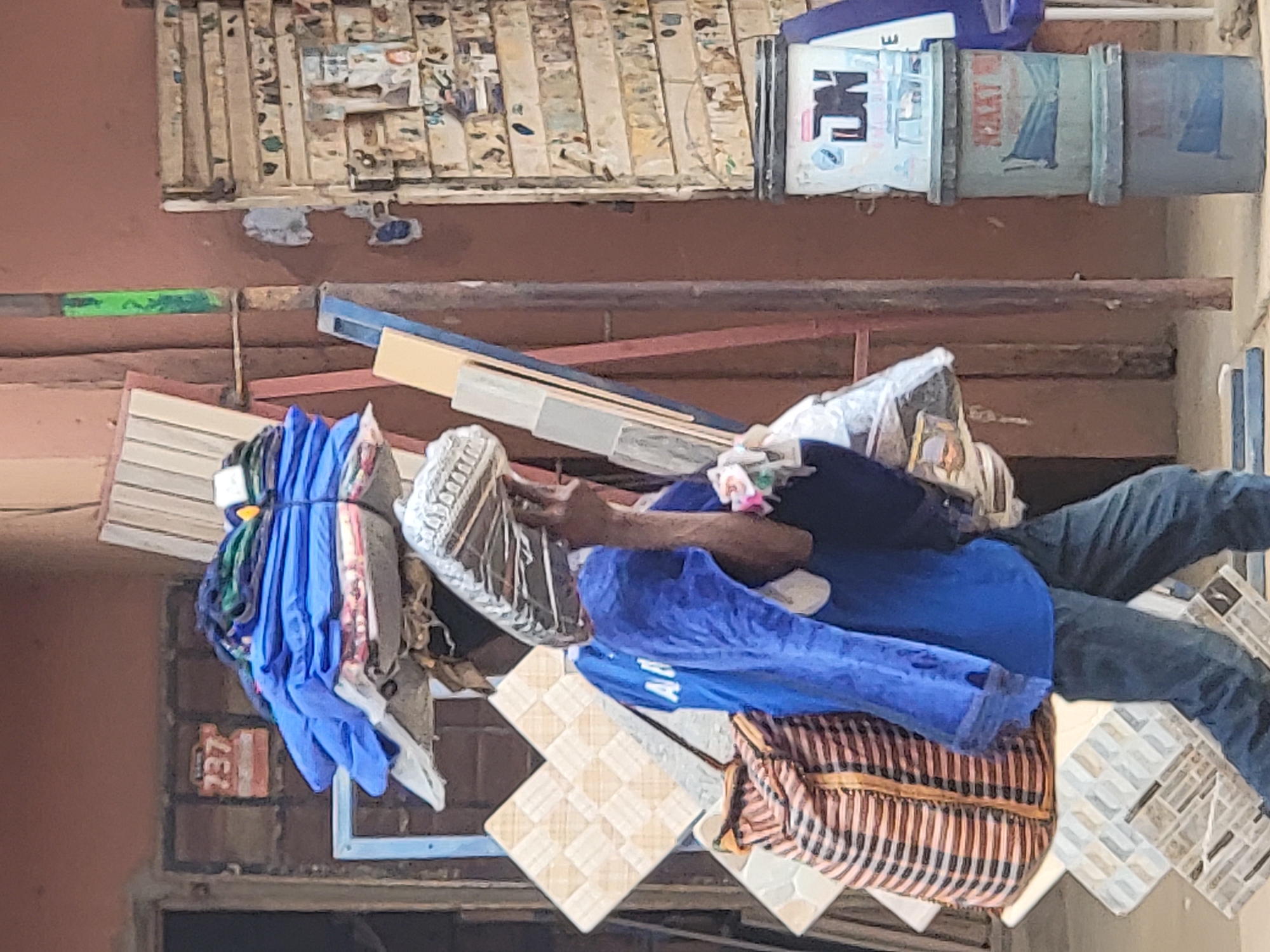 A man wearing a blue shirt and jeans is sitting amidst a collection of objects, including a bag, tiles, and bundled clothing. The location is Maiburiji, Nigeria, indicated by "MAI" visible on an object above. The background reveals a brick wall with a painted number "737" and corrugated metal sheets. A weathered shelf holding cylindrical containers is present above the man.