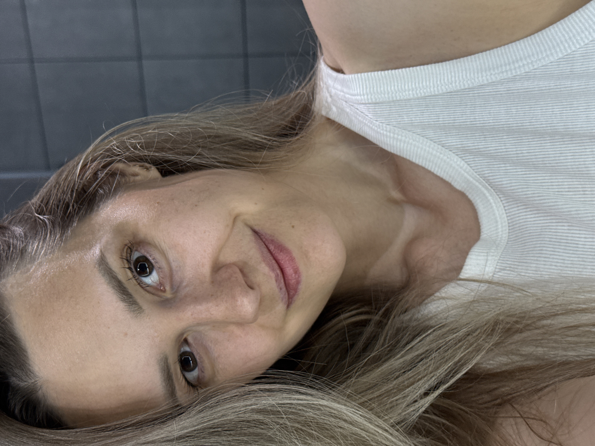 A close-up, slightly angled portrait shows a woman with fair skin, freckles, and light brown hair with blonde highlights. She is wearing a white ribbed tank top. She is looking directly at the camera with a slight smile. The background appears to be a wall tiled with dark gray rectangular tiles. There is no information available in the image about the location in Chaikovsky, Russia.