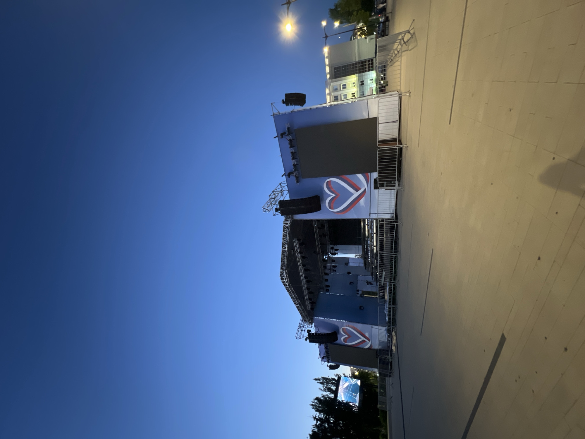 The image shows an outdoor stage set up in a paved plaza, likely for an event. The stage is a large, multi-level structure with a roof and lighting rigs. Two large screens are visible on either side of the stage, and a large heart symbol with red, white, and blue colors is painted on the backdrop of the stage. Metal barricades are set up in front of the stage, suggesting crowd control measures. In the background, there are trees, a street lamp, and a multi-story building with lit windows. The sky is a clear, deep blue, indicating it is either early morning or late evening. The overall impression is of a prepared venue awaiting a performance or event.
Based on the presence of the tricolor heart symbol, which strongly resembles the Russian flag, and the context of outdoor event staging, this scene is likely located in Russia. Without further information or context, pinpointing the exact location within the city of Izhevsk, Russia, is not possible from the image alone. However, the setup suggests a public gathering space, such as a central square or park, commonly used for concerts or festivals.