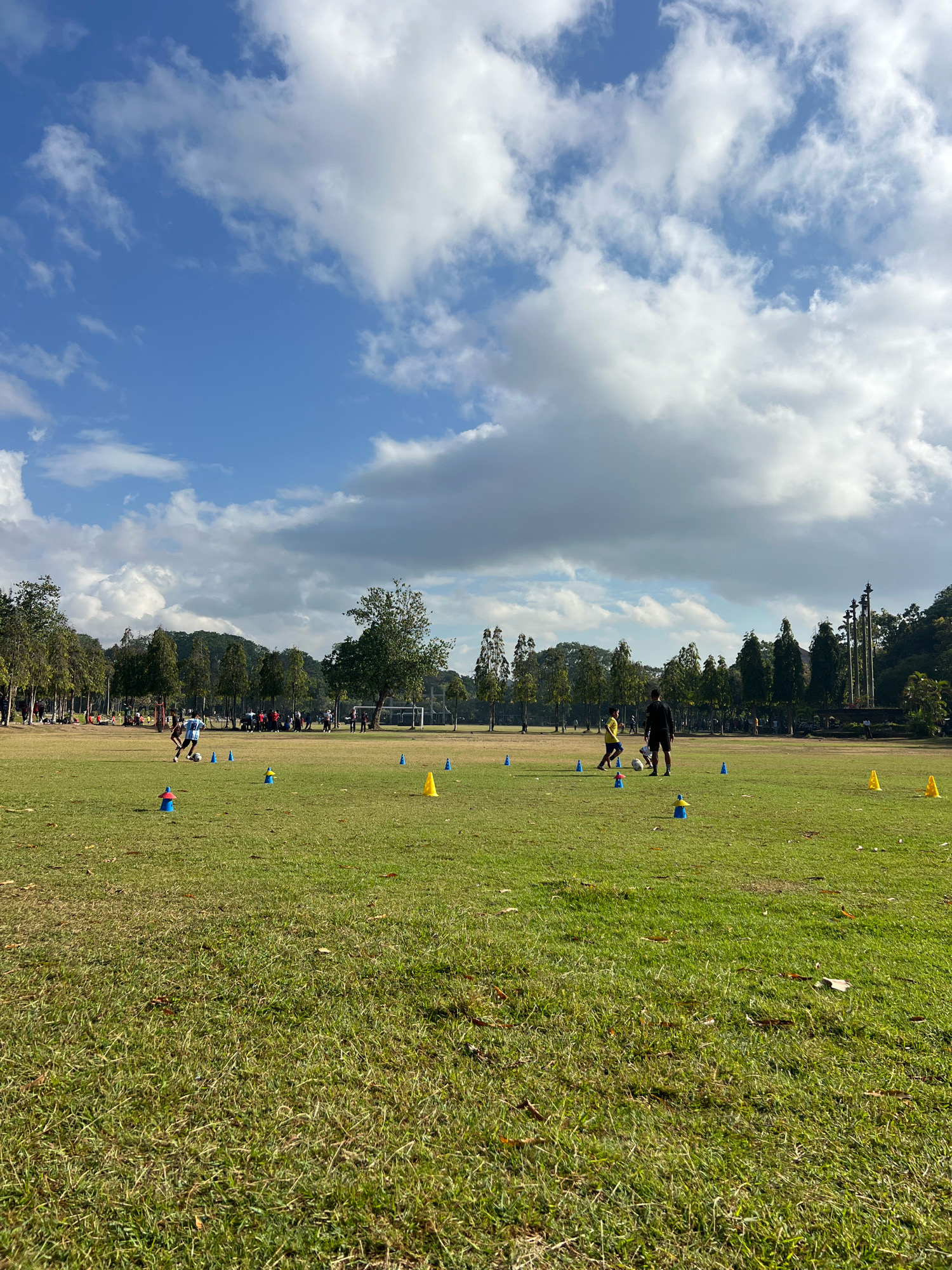 The image captures a wide view of a grassy field on a partly cloudy day, with a line of trees and some distant structures forming the background. In the foreground, a patch of grass is visible, with some fallen leaves scattered on it. The mid-ground shows a sports field with several people, including children and adults, engaged in what appears to be a soccer training session. There are multiple blue and yellow cones placed on the field, likely for drills. Two individuals are actively playing soccer with a ball, one wearing a blue and white striped shirt and the other a yellow shirt. Another adult, wearing a black shirt, is also present on the field. In the background, a line of trees borders the field, and beyond that, more trees and hills are visible under a sky with scattered clouds. There are also some distant structures and a soccer goal visible on the right side of the field. The scene suggests an outdoor sports or recreational activity taking place in a park or open space within Denpasar, Indonesia.