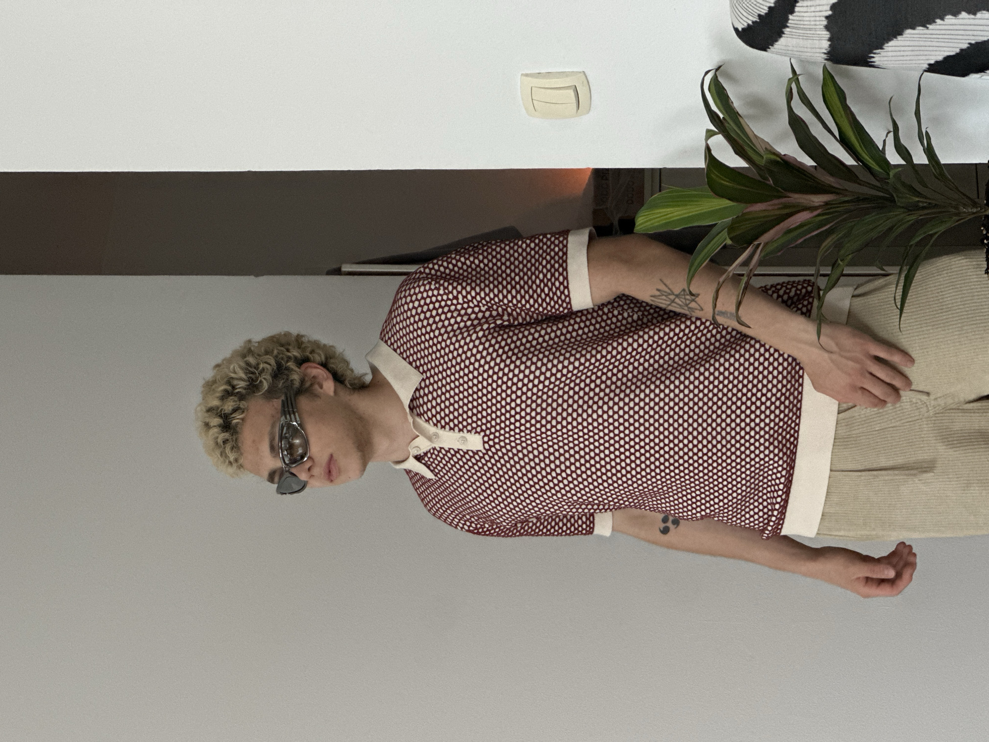 A full-length, slightly off-center portrait features a person with curly blonde hair and unique sunglasses, standing in an indoor setting. The person is wearing a red and white patterned short-sleeved polo shirt with a cream-colored collar, placket, and sleeve cuffs. Their light-colored (possibly beige or cream) trousers appear to have a corduroy or ribbed texture. Visible tattoos include a geometric or abstract design on the left forearm and a smaller symbol on the right forearm, possibly a semicolon. The sunglasses are distinctive, featuring a metallic, multi-faceted frame design.
The background consists of a plain, light grey wall. To the right of the person, a white shelf or surface holds a houseplant with long, broad green leaves that have hints of pinkish-red. Above this shelf, on the wall, there is a cream-colored electrical switch or outlet. A horizontal dark brown stripe or panel is visible on the wall behind the person's midsection, separating the lighter grey lower wall from a white upper wall. In the upper right corner, a glimpse of a black and white patterned object, possibly a cushion or fabric, can be seen. The lighting is soft and even, suggesting an indoor environment, with no discernible external light source or time of day. There is no visible text in the image.
