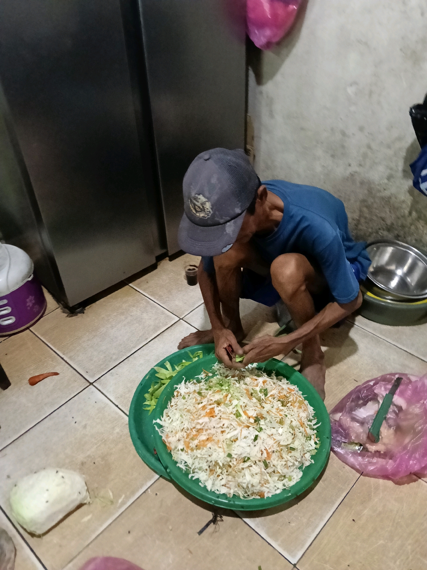 In the city of Belimbing, Indonesia, a man is crouched on a tiled floor next to a large green bowl filled with shredded vegetables, likely cabbage and carrots. He is wearing a blue t-shirt and a dark baseball cap. His hands are focused on chopping more vegetables. To the right of the bowl, a pink plastic bag is visible, containing some food items. In the background, a large, dark refrigerator stands against a light-colored, textured wall. The overall scene appears to be an indoor domestic setting, possibly a kitchen or an open-air area of a home.
