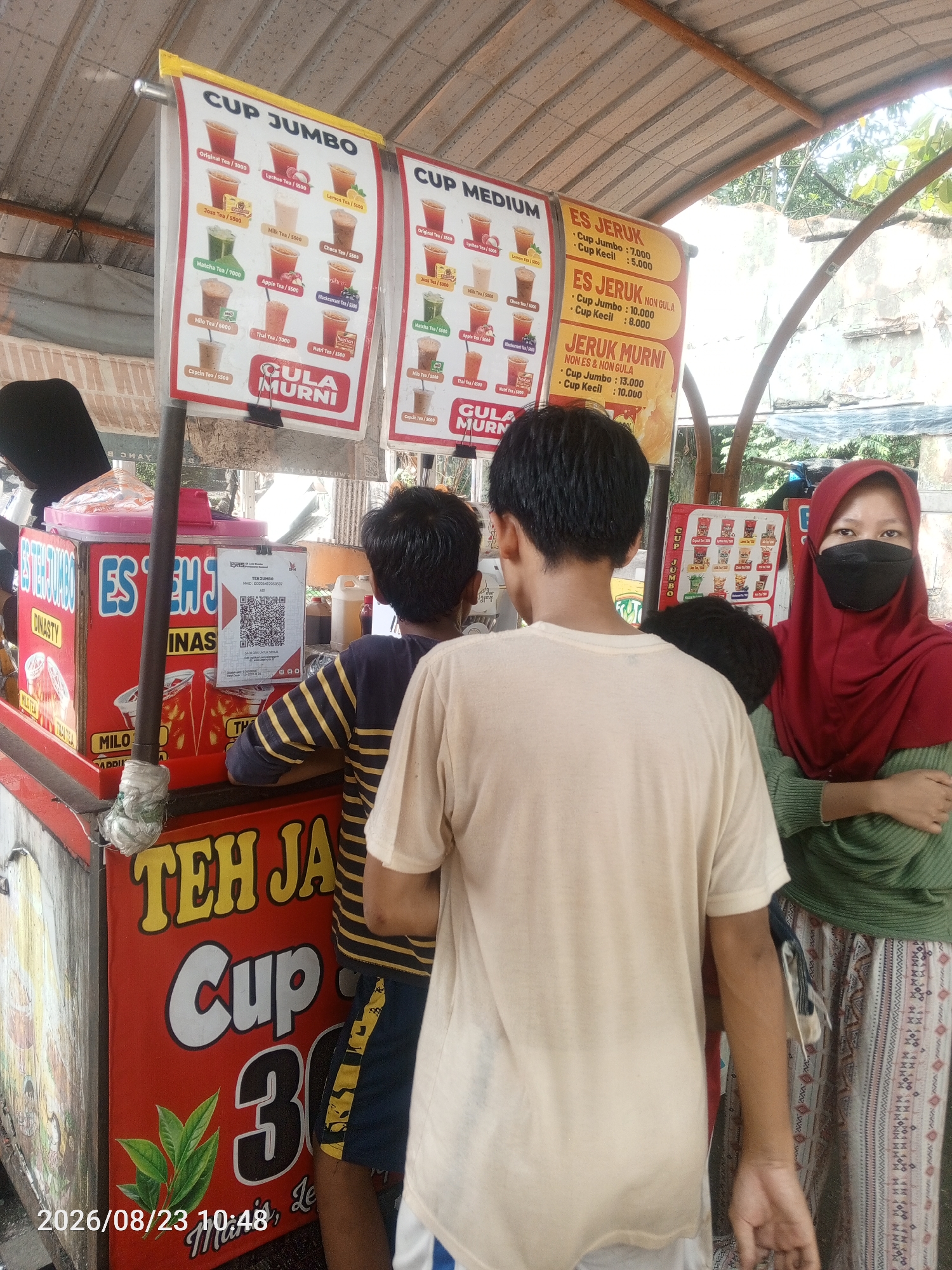 In Depok, Indonesia, at a street-side beverage stall, a woman wearing a maroon hijab and a black face mask stands on the right, her arms crossed. To her left, a young man in a cream-colored t-shirt stands with his back to the camera, his right arm extended. Further left, another young person in a striped shirt is visible. The stall itself is red with yellow lettering that says "TEH JAL". A menu board above the stall lists various tea and juice options, including "CUP JUMBO" and "CUP MEDIUM" with prices. Another sign, also red with yellow text, advertises "TEH JAL Cup 30". A QR code is visible on the stall for payment. The scene appears to be outdoors under a corrugated metal roof, with greenery in the background. The timestamp "2026/08/23 10:48" is visible in the bottom left corner.