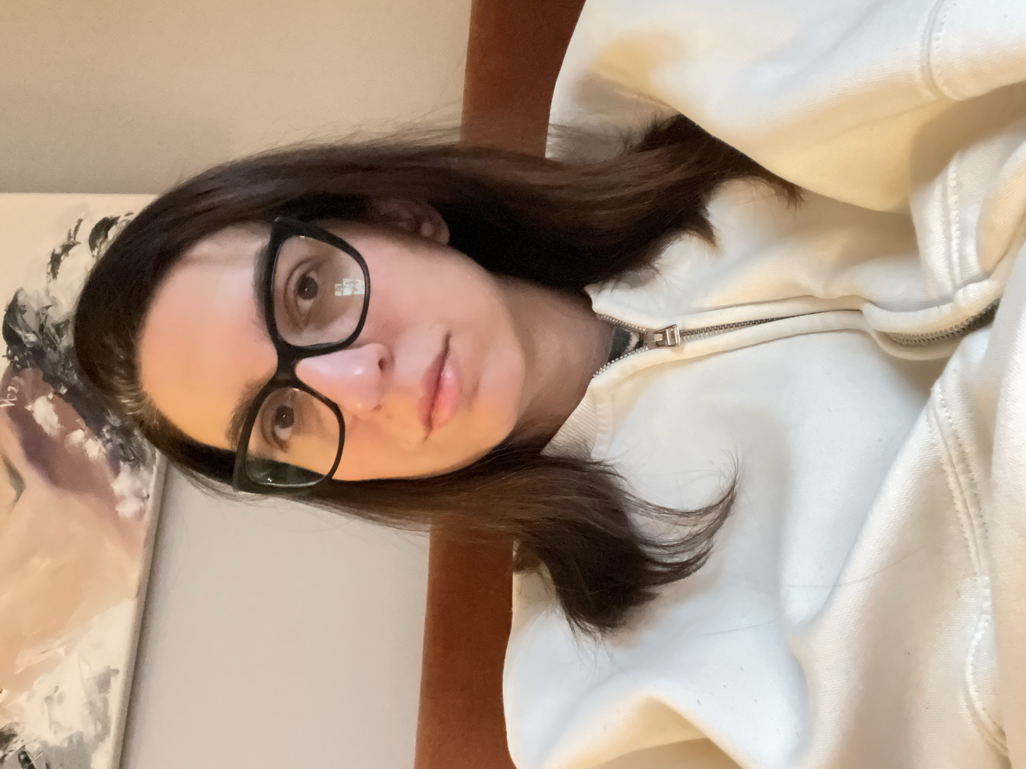 A woman with brown hair and glasses is wearing a white zip-up sweatshirt. There is a painting visible in the background. The location is unknown; there is no evidence in the image to suggest the city of Sofia, Bulgaria.
