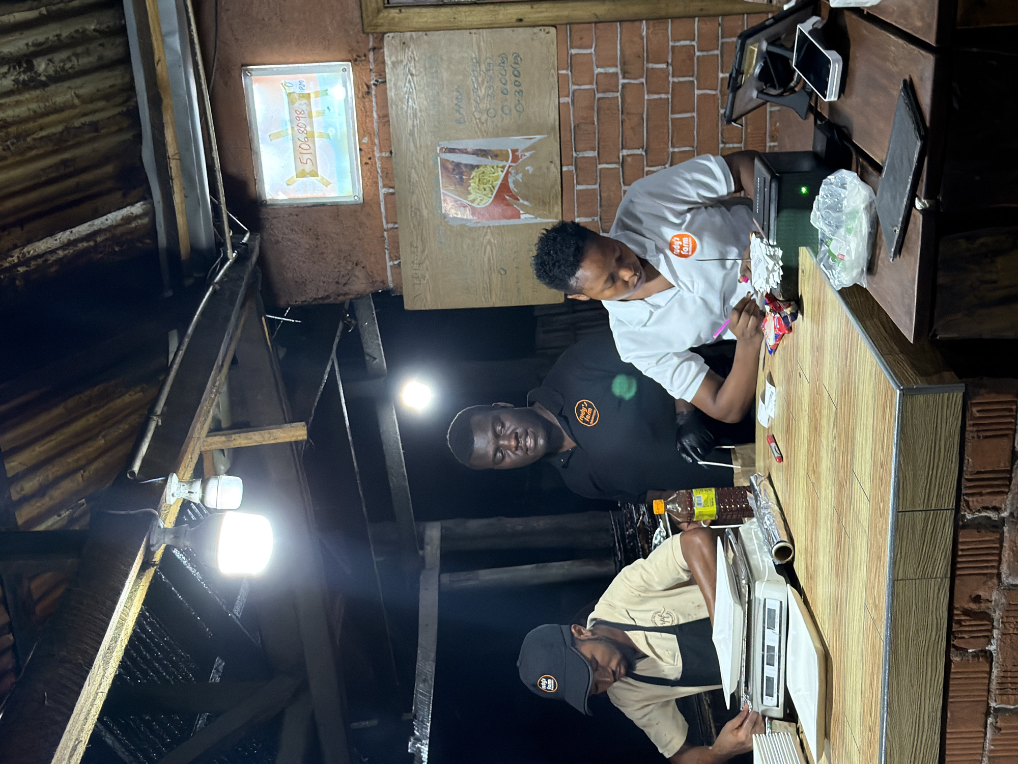 In Dar es Salaam, Tanzania, three individuals are working behind a wooden counter in what appears to be an outdoor food stall or small restaurant. The man on the left, wearing a black cap and a tan apron over a light-colored shirt, is focused on a digital scale with white plates. To his right, a man in a black polo shirt with an orange logo is looking forward with a serious expression. The person on the right, wearing a white polo shirt with a similar orange logo, is writing on a stack of papers with a pink pen, with a card reader and a tablet nearby. The environment suggests a rustic setting with a corrugated metal roof supported by wooden beams. A bright LED light illuminates the workspace. There is a bottle of sauce, foil-wrapped items, and a notepad on the counter. A handwritten menu or price list is visible on a wooden board on the brick wall behind the counter. A sign with a phone number is also displayed on the wall.