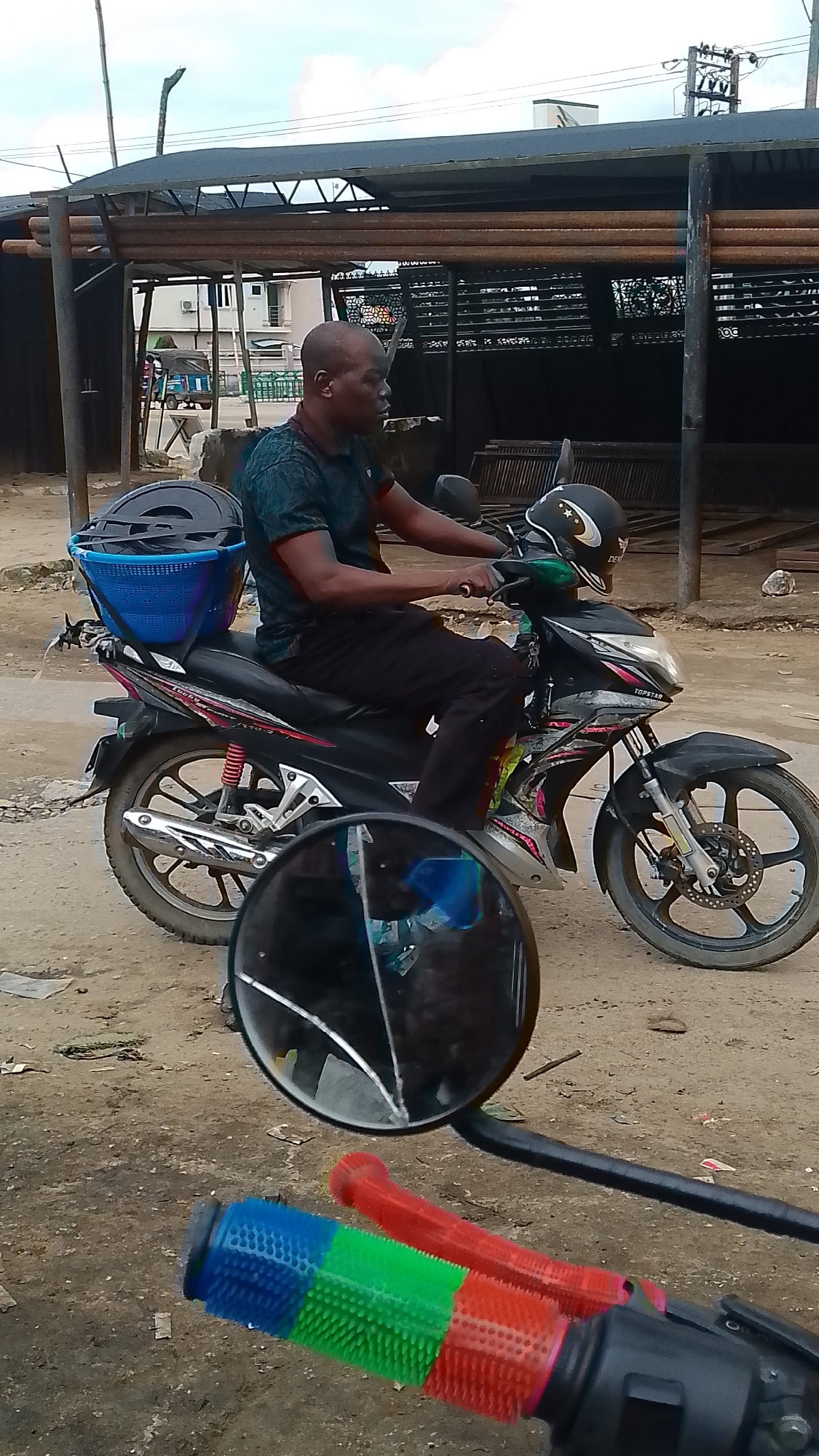 A man is riding a black motorcycle with a blue basket strapped to the back. The motorcycle has red and silver accents. The man is wearing a dark t-shirt and black pants, and a black helmet with a star design. The motorcycle has a "TOPSTAR" sticker on the front. In the foreground, the handlebars of another motorcycle are visible, with bright blue, green, and red grips. The scene appears to be in a dusty, outdoor urban environment, likely in Warri, Nigeria, with some weathered structures and debris in the background. A broken mirror is positioned in the lower part of the image, reflecting the sky and possibly the rider's face.