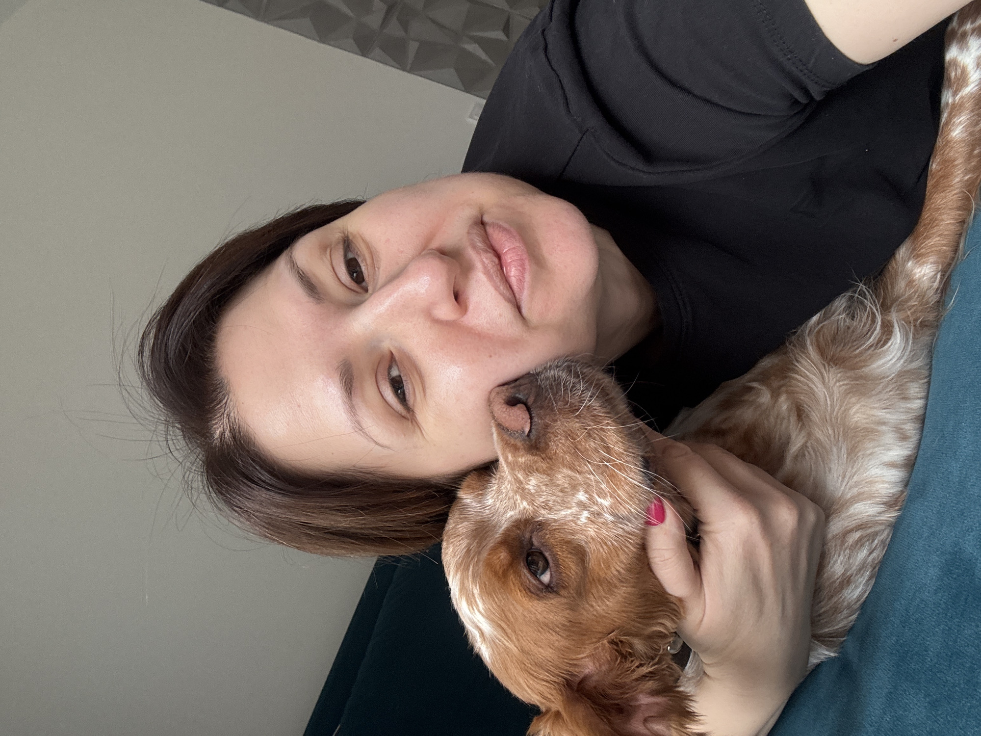 A medium-shot photograph shows a woman and a dog indoors. The woman, fair-skinned with brown hair, is wearing a black shirt. Her face is close to the dog's head. The dog is light brown and white with floppy ears. The woman's hand is visible, holding the dog near its neck. The background is a combination of a textured white wall, a greyish-white wall, and a blue-green cushion or piece of furniture. The location is indicated as Sofia, Bulgaria, but there is no explicit visual evidence within the image to confirm this.