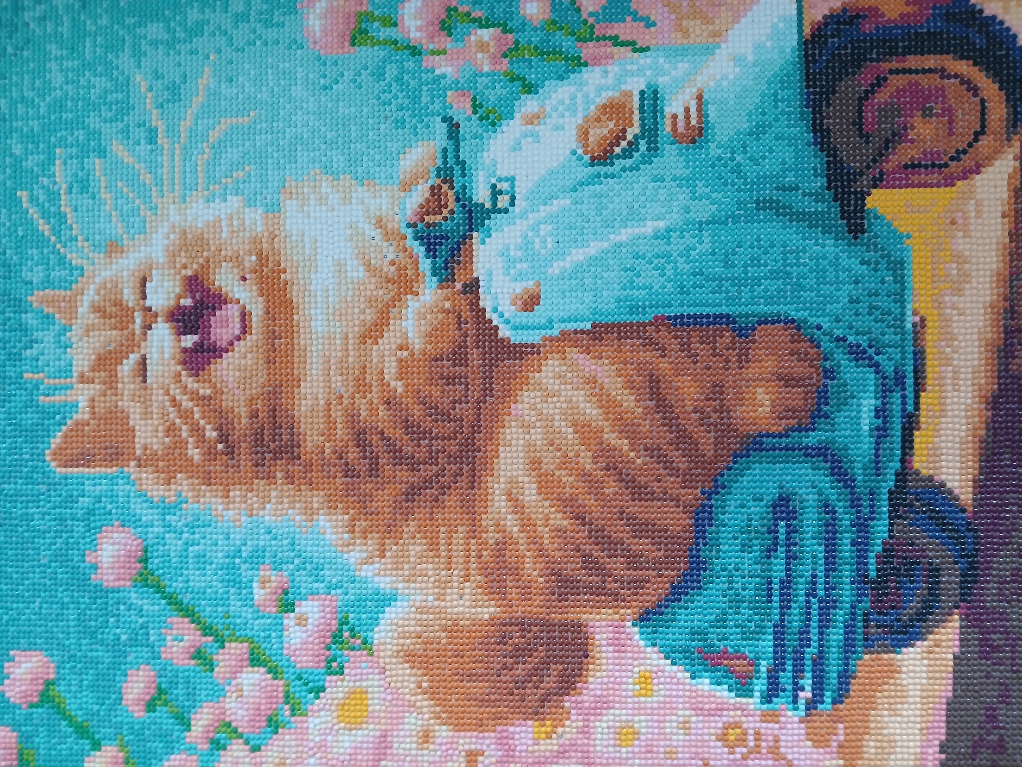 This image is a close-up of a diamond painting featuring an orange cat riding a light blue scooter. The cat has its mouth open in a cheerful expression, with its tongue sticking out. It is surrounded by pink and white flowers against a turquoise background. The scooter has a vintage design, with chrome accents and a visible wheel. The diamond painting style creates a textured, mosaic-like appearance. There is no discernible location within the city of Korenovsk, Russia, depicted in this artwork.