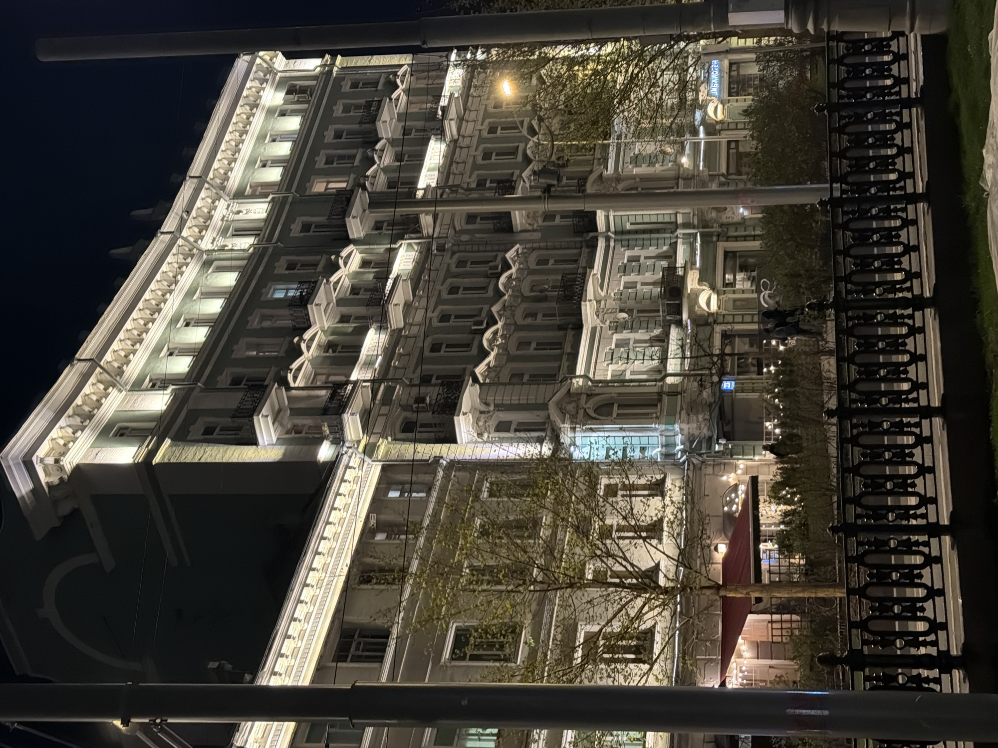 This night-time photograph captures a grand, multi-story building in Moscow, Russia, illuminated by external lighting. The building showcases classical architectural elements, including elaborate cornices, decorative window frames, and small wrought-iron balconies. Its light-colored facade (likely grey or beige) is accentuated by floodlights, highlighting the intricate details of its structure.
In front of the building, trees with bare branches suggest the season is late autumn or winter. A decorative black wrought-iron fence runs along the lower right of the image, separating the viewer from the street level. The ground floor houses various establishments, indicated by warmly lit windows and, on the right side, a prominent red awning covering an outdoor seating area adorned with string lights. Two dark, indistinct figures are faintly visible behind the fence, near the red awning.
The scene is set at night, with a dark sky dominating the upper left portion of the image, contrasting with the abundant artificial light emanating from the building and street fixtures. Visible text includes "ШЕКСПИР" (SHAKESPEARE) on a sign, possibly indicating a business like a restaurant or bar. Another circular sign on the ground floor is present but its text is unreadable. The overall ambiance is one of serene urban elegance, characteristic of a historical city center after dark.