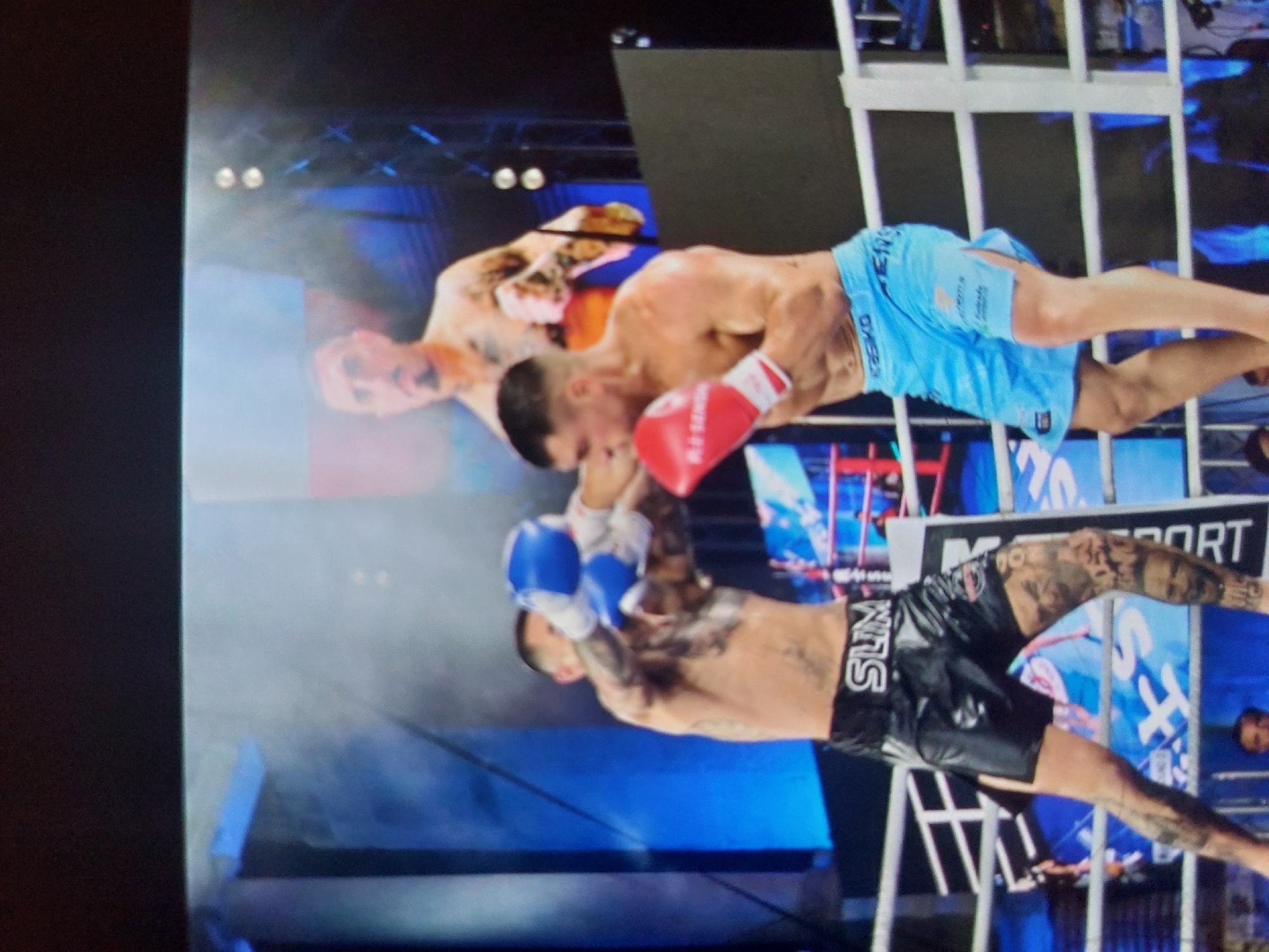 The image captures an intense moment during a combat sports event, likely boxing or kickboxing, set within an indoor arena. Two male fighters are centrally positioned in the ring, engaged in a dynamic exchange.
The fighter on the right wears light blue shorts with some text (partially legible) and bright red boxing gloves. He has extensive tattoos covering his arms and torso. His gaze is fixed, and he appears to be in the process of throwing or having just thrown a punch towards his opponent.
The fighter on the left wears black shorts with the white text "SLIM" clearly visible on the waistband, paired with blue boxing gloves. He also has numerous tattoos on his arms and legs. His body language suggests he is bracing for impact or preparing a defensive maneuver.
White vertical structures, resembling the ropes or bars of a fighting cage/ring, frame the fighters and extend across the scene. The background features bright stage lighting, creating a dramatic atmosphere typical of such events. Large screens or banners display partially visible graphics, including the text "M SPORT". Blurred figures in the distance hint at an audience, while other individuals, like a man with a beard partially visible in the background, watch the action. The event unfolds under artificial illumination, indicating it is likely evening or night. The location context provided is Tsarevo, Bulgaria. The image itself appears to be a photograph taken of a screen, contributing to a slight blurriness.