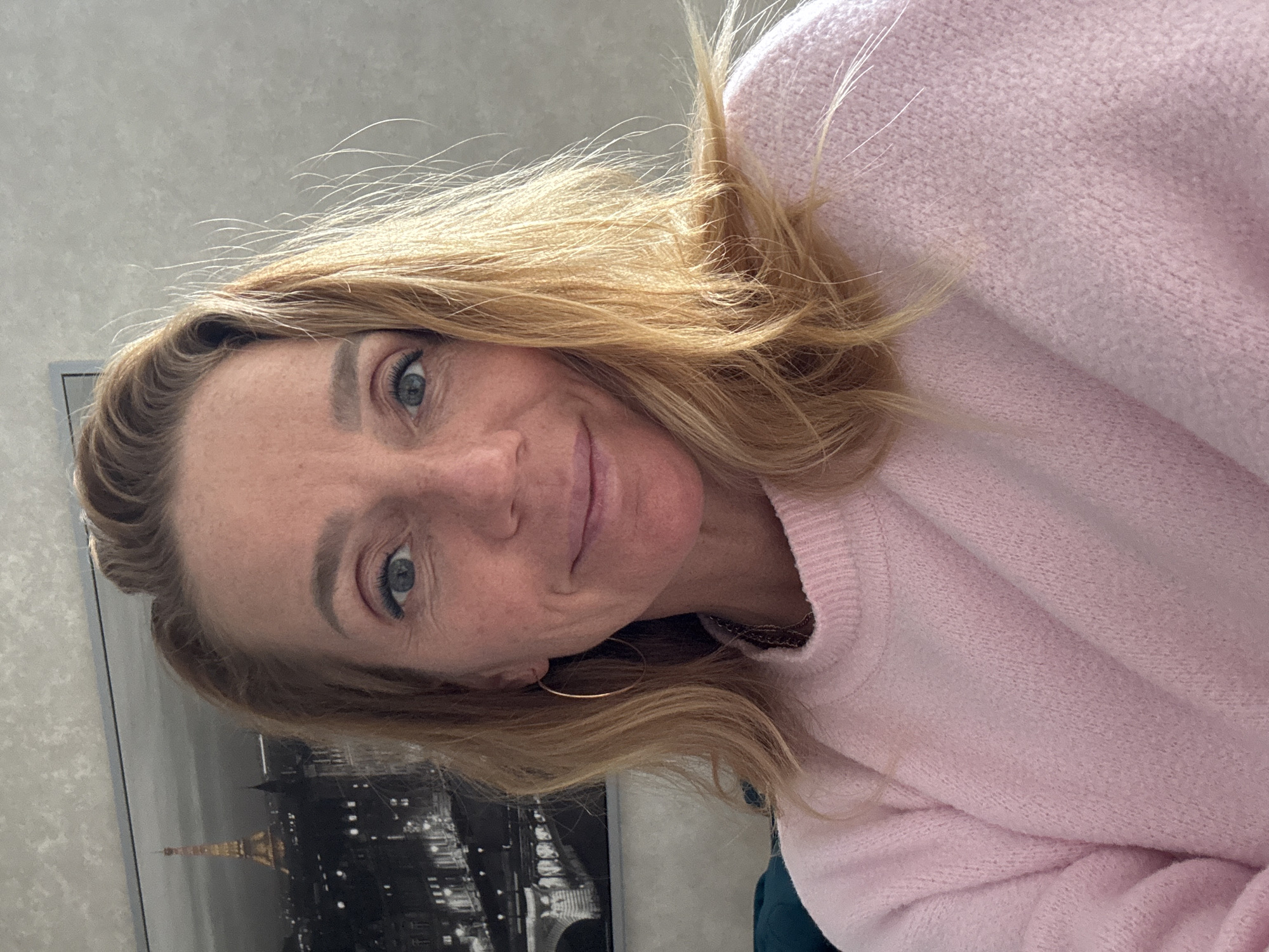 A woman with blond hair and blue eyes is wearing a pink sweater. She is smiling slightly. Behind her is a framed picture of Paris, France, featuring the Eiffel Tower. The location is unspecified, and there is no indication of Saint Petersburg, Russia.