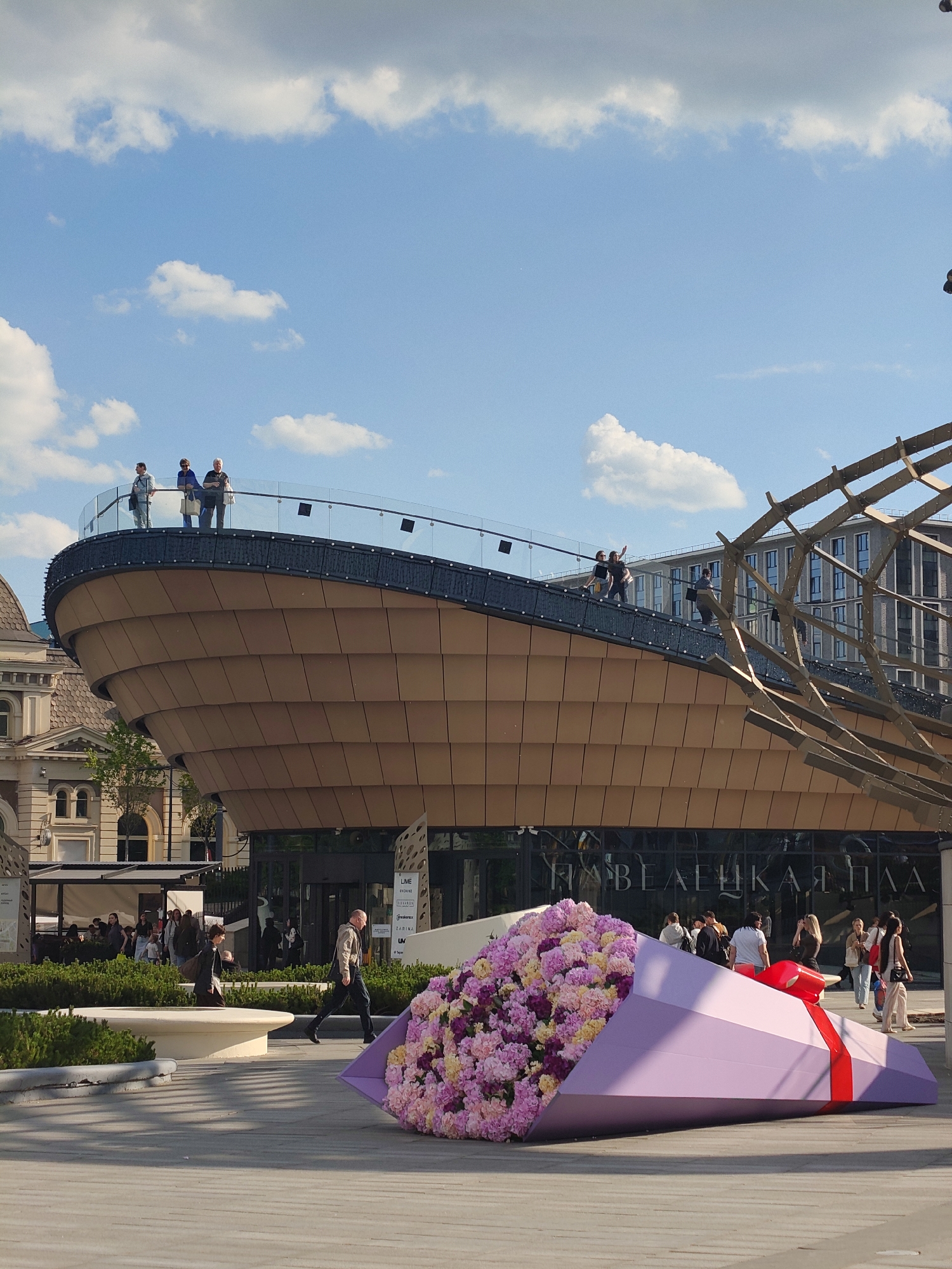 The image captures a vibrant urban plaza under a bright blue sky with scattered white clouds. In the foreground, a prominent artistic installation resembling an oversized bouquet of artificial flowers lies on the paved ground. The bouquet features a dense cluster of pink, purple, and yellow blooms, encased in a geometric lavender-colored wrap with a striking red bow.
The central subject is a distinctive modern building with a curved, multi-tiered brown facade, topped by an accessible deck featuring a glass railing. Several individuals are visible on this upper deck, standing and observing the surroundings. More people are scattered throughout the plaza at ground level, walking casually or congregating near the building's entrance. To the right, an architectural element composed of dark, angular beams forms an open, skeletal structure.
The bright sunlight suggests it is daytime, casting clear shadows across the plaza. On the reflective glass facade of the main building, the Cyrillic text "П А В Е Л Е Ц К А Я П Л А" is clearly visible, suggesting the location is "Paveletskaya Plaza." Smaller text, including "LIME" and "ZARINA," can also be seen near the building's entrance. The scene conveys a relaxed, public atmosphere in a contemporary urban setting.
