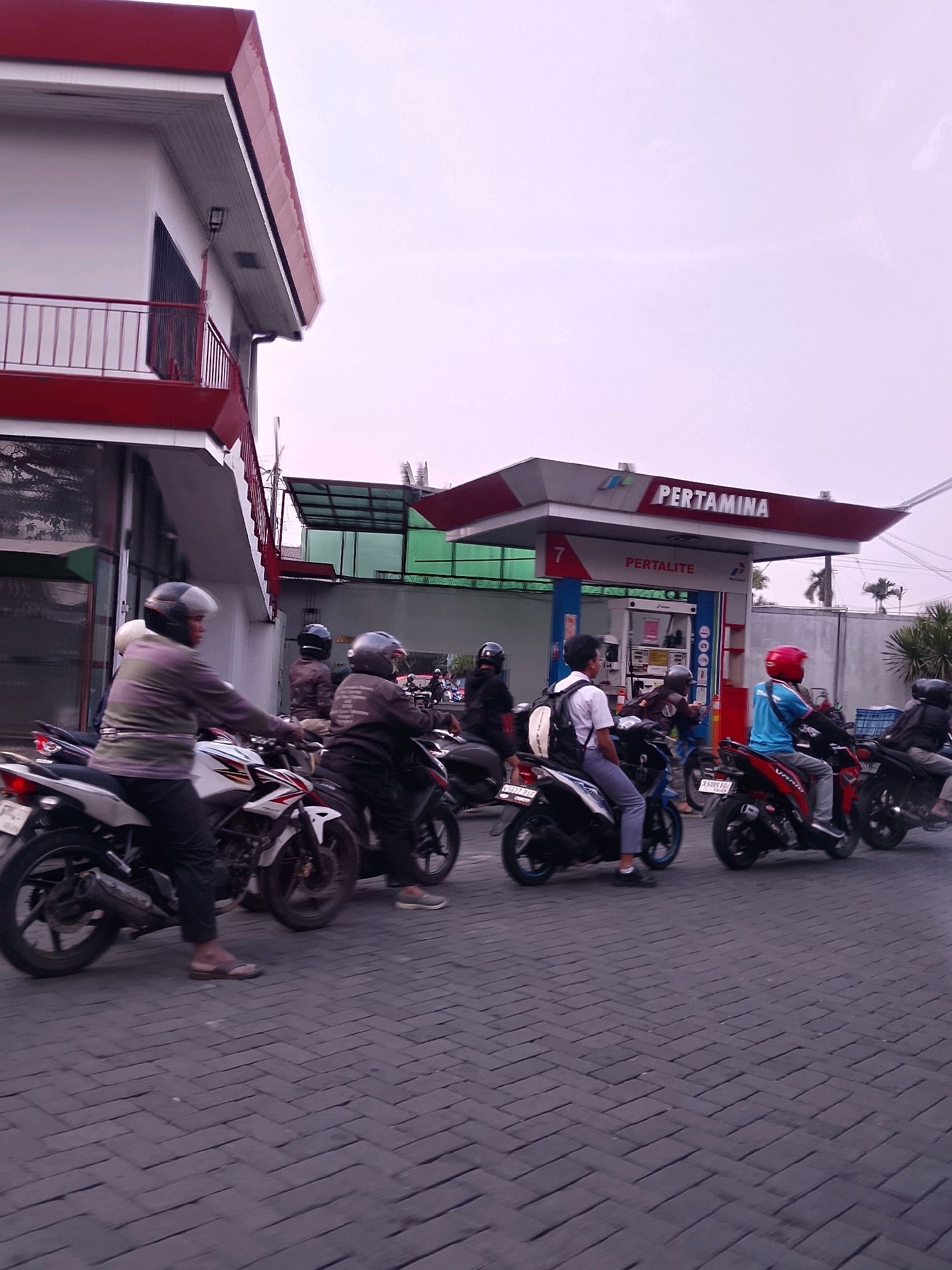 This is a vertical, medium-angle, outdoor photograph of a Pertamina gas station in Malang, Indonesia. The sky is overcast. In the foreground, several people on motorcycles are lined up, appearing to be waiting for service. The Pertamina station canopy is visible in the background with the Pertamina logo prominently displayed. The ground is paved with dark gray brick pavers arranged in a herringbone pattern. The lighting suggests it is daytime, possibly late afternoon.