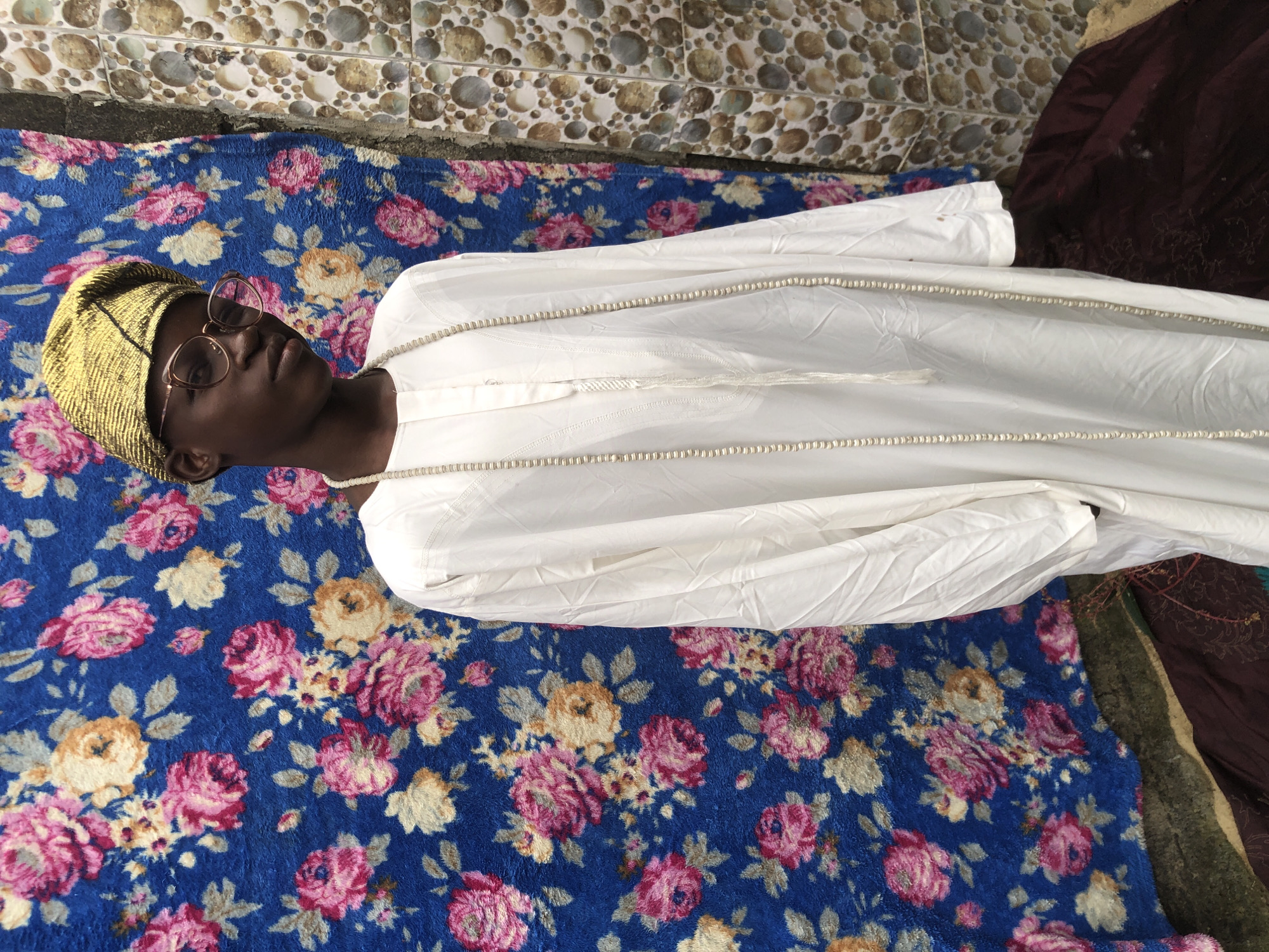 A young Black male stands in front of a floral patterned blue rug and a tiled wall. He wears a gold beaded cap, clear-rimmed glasses, and a white robe with a beaded necklace. He is looking slightly to his left. The setting appears to be outdoors, possibly in Ado, Nigeria, with a tiled wall on the right side of the frame and a brown cloth at the bottom.