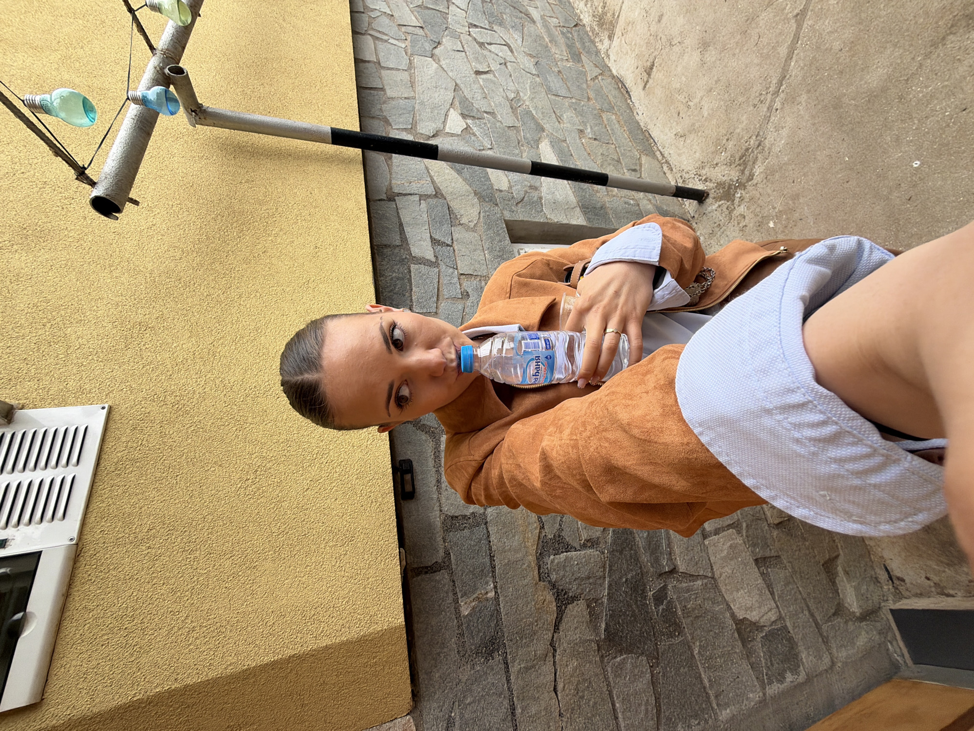 The image shows a woman drinking from a water bottle while standing outside in what appears to be an urban environment. She is wearing a brown jacket and a light blue shirt. The background includes a yellow wall, a stone walkway or street, and a metal pipe structure with attached bottles. The water bottle label has Cyrillic characters, and given the location provided, it is likely in Bulgarian. The location is specified as the city of Buhovo, Bulgaria.