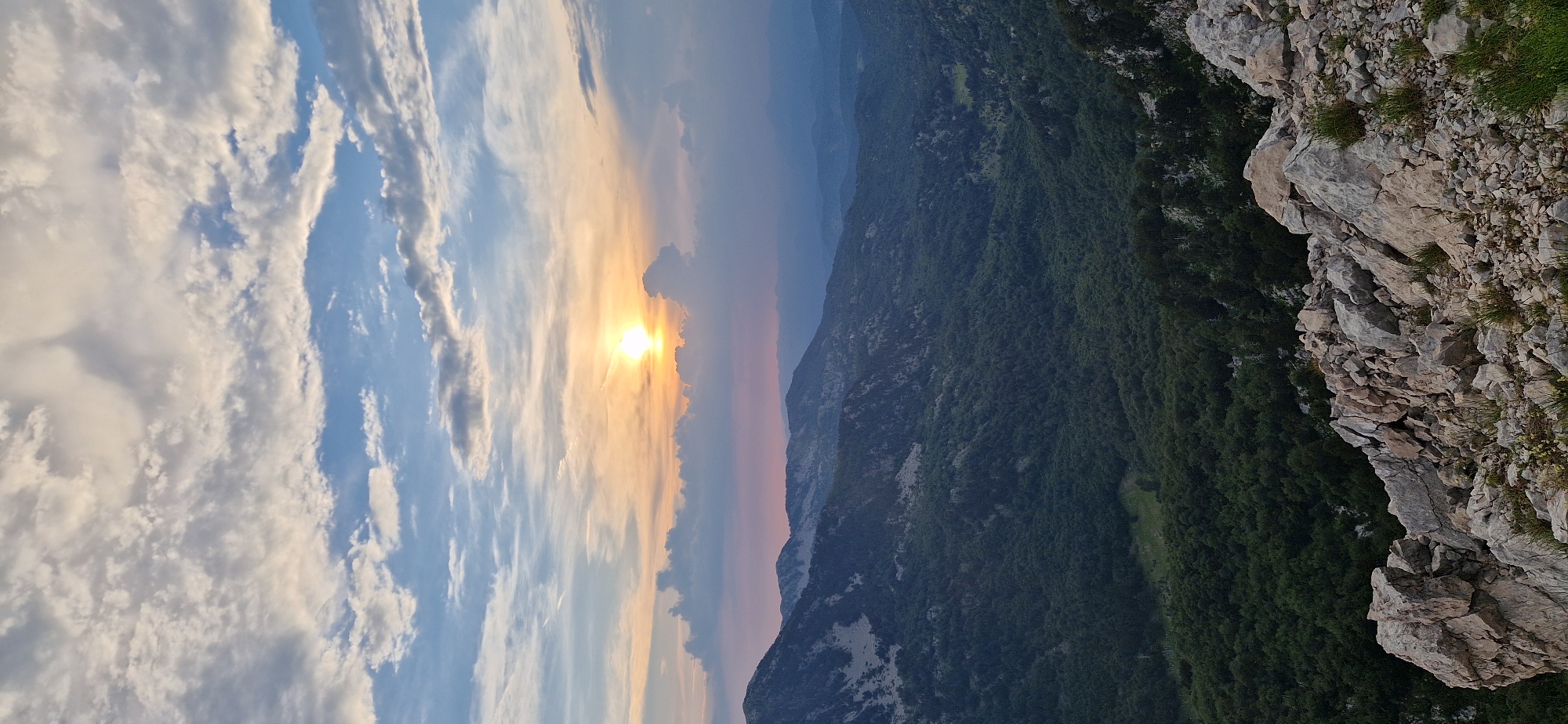 An aerial view captures a sunset over a mountainous landscape. The sun, a bright orb with visible rays, is positioned centrally in the sky, casting a warm orange and yellow glow that blends into softer pinks and purples towards the horizon. Clouds, varied in texture and density, drift across the sky, some catching the sunlight and appearing illuminated.
Below the sky, the terrain transitions into a series of rugged mountains. The closest mountains in the foreground are rocky and arid, with sharp, angular rock formations. These give way to a dense forest that carpets the slopes and valleys of the mid-ground and background. The trees are a deep, rich green, suggesting a thick, verdant growth. Further in the distance, successive mountain ranges recede into a hazy, muted blue, indicating atmospheric perspective. The overall impression is one of a remote, natural environment bathed in the soft light of dusk. The location is identified as Ubla, Bosnia and Herzegovina.