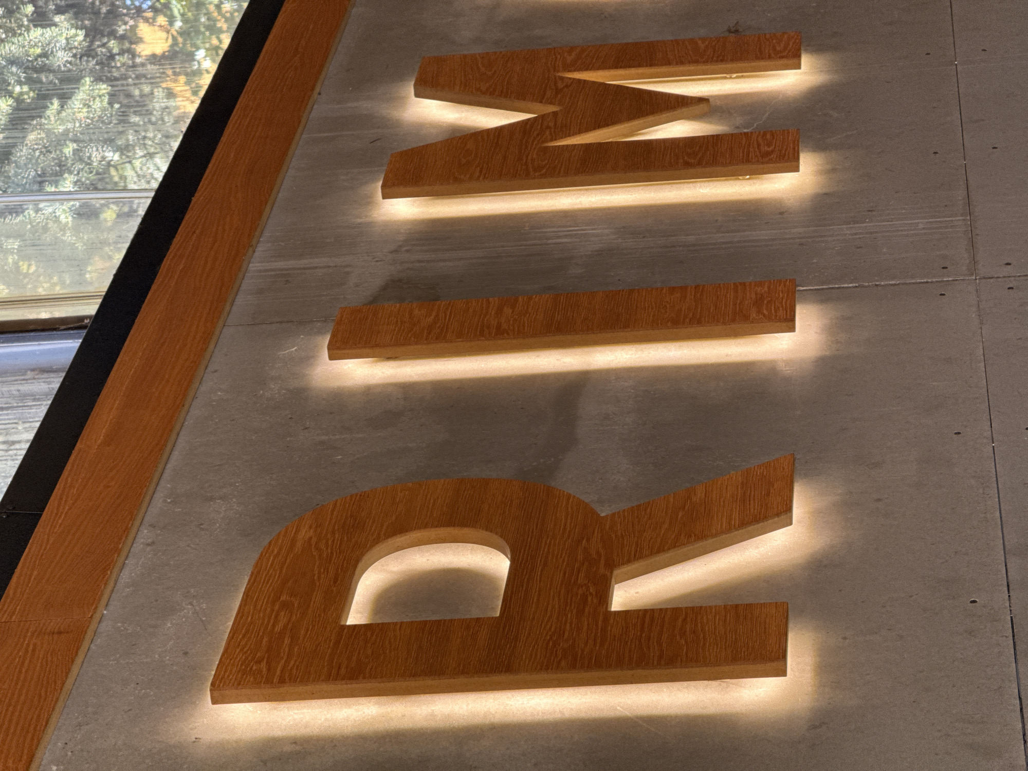 The image shows a close-up of large, wooden, illuminated letters mounted on a concrete wall. The letters spell out "RIM". The wood has a visible grain and is a warm, medium brown color. The letters are lit from behind, casting a soft, warm glow on the concrete surface. Above the letters, there is a wooden beam and a black strip that appears to be part of the building's facade. A window with trees visible outside is partially visible at the top right of the image. The scene appears to be an interior or exterior sign. There are no people or animals in the image. The location in the city of Shumen, Bulgaria is not discernible from the image itself.