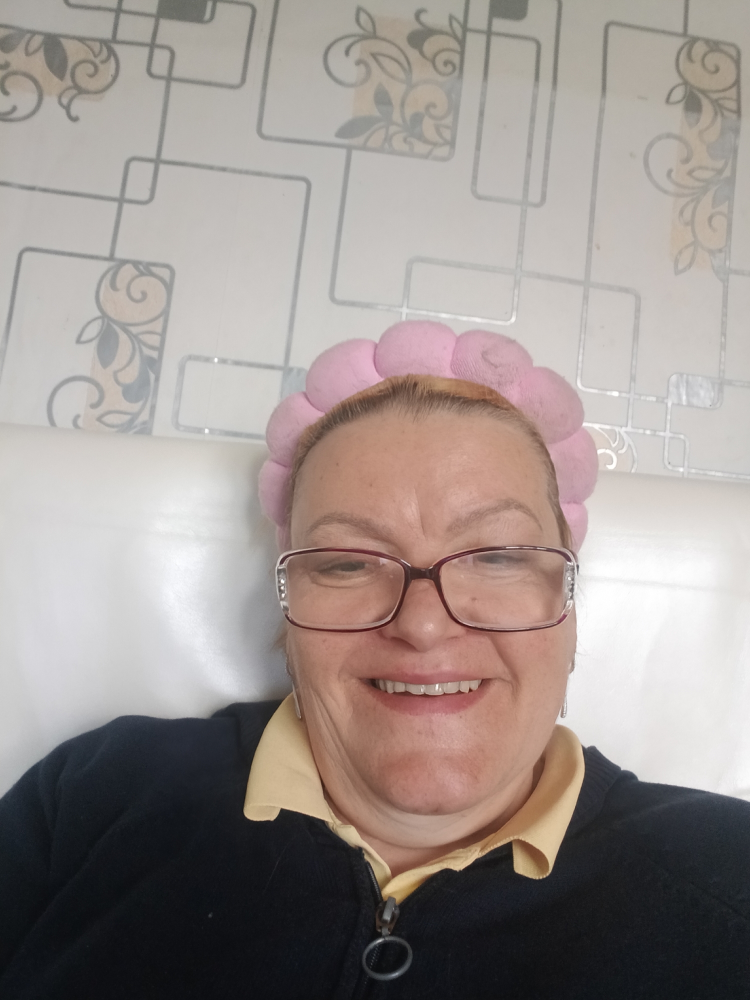A middle-aged woman with blonde hair and glasses is smiling at the camera. She is wearing a pink, fluffy headband and a black jacket over a yellow collared shirt. She appears to be sitting against a white headboard or sofa. The background is a white wallpaper with a geometric pattern and floral designs. The image may have been taken indoors. There is no information in the image to confirm it was taken in Красный Маяк, Russia.