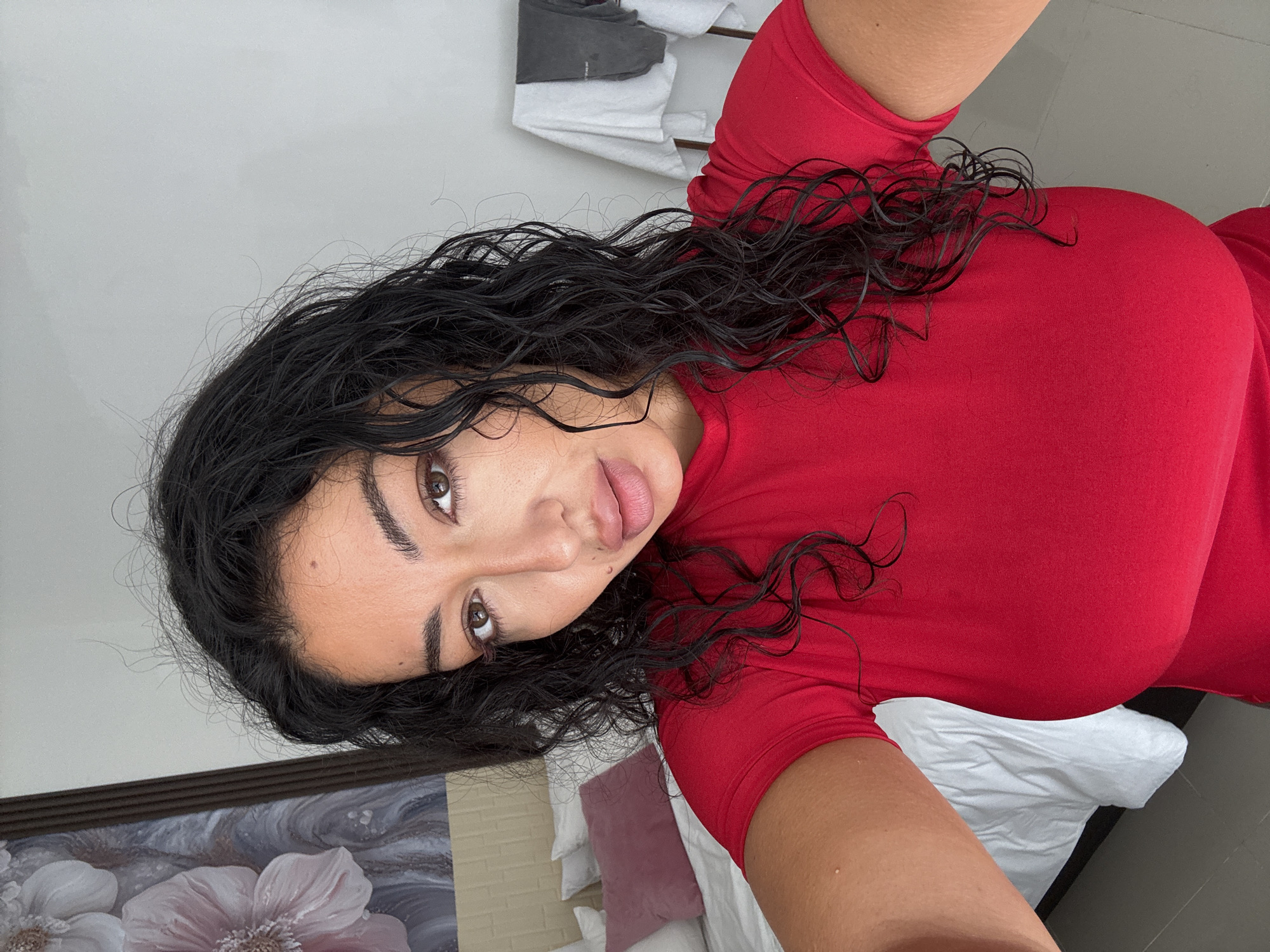 A selfie taken indoors in Kuta Selatan, Indonesia, shows a woman with dark, wavy hair wearing a red, short-sleeved shirt. Her hair appears wet. Behind her is a bed with white linens and several pillows in shades of white and pink. Part of a framed floral painting is visible in the lower left corner. Clothes are hanging on the wall in the background.