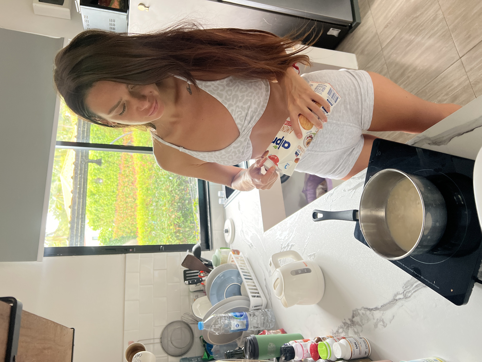 A young woman stands in a well-lit kitchen located in the city of บ้านบางรักษ์, Thailand, actively engaged in food preparation. She possesses long, dark brown hair and is attired in a light-colored, leopard-print sports bra and light grey shorts. She is holding an Alpro almond milk carton, seemingly pouring its contents into a metal saucepan positioned on a black portable induction cooktop, which contains a light-colored liquid. The kitchen features white countertops with a grey marble-like pattern and a white subway tile backsplash. To the left of the scene, various kitchen items are arranged, including a white Tefal electric kettle, a dish drying rack with blue, grey, and white plates alongside a "Crystal" brand water bottle, and an assortment of supplement bottles, notably one labeled "CALIFORNIAN GOLD NMN". A large window in the background reveals lush green tropical foliage and parts of a building roof, with a grey roller blind partially drawn.