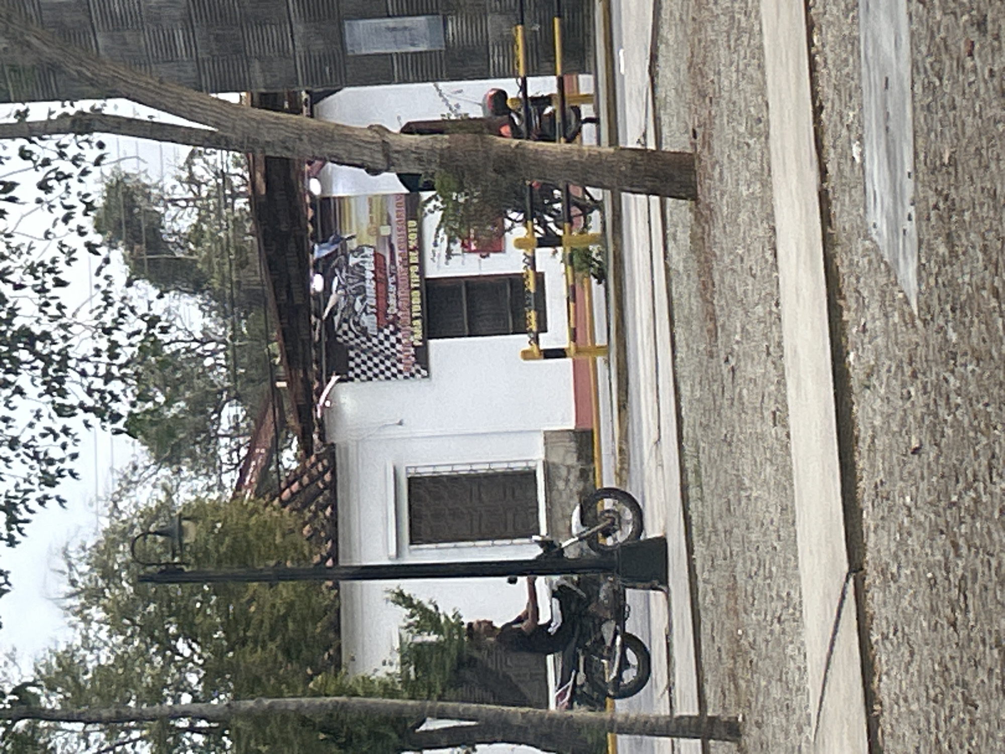 A side profile of a man riding a white motorcycle down a paved road. The man is wearing a dark t-shirt and dark pants. The motorcycle has a dark seat and silver accents. Behind the motorcycle, there is a white building with a red tiled roof. A banner is displayed on the building with checkered flags and text. A street light is visible to the left of the motorcycle. The environment appears to be a street in Santa Ana, Venezuela, with trees and foliage in the background.