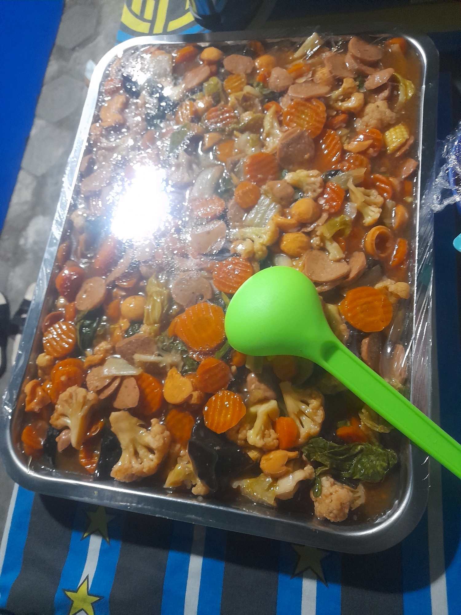 In Mlaras, Indonesia, a large rectangular metal tray filled with a dish of stir-fried vegetables and sliced sausages is presented. The dish is covered with plastic wrap, and a bright green ladle rests on top of the food, partially submerged in the sauce. The tray is positioned on a surface with a striped blue and gray pattern, and a yellow star emblem is visible in the lower left corner. The overall lighting suggests an indoor environment, possibly a home or catering setup.