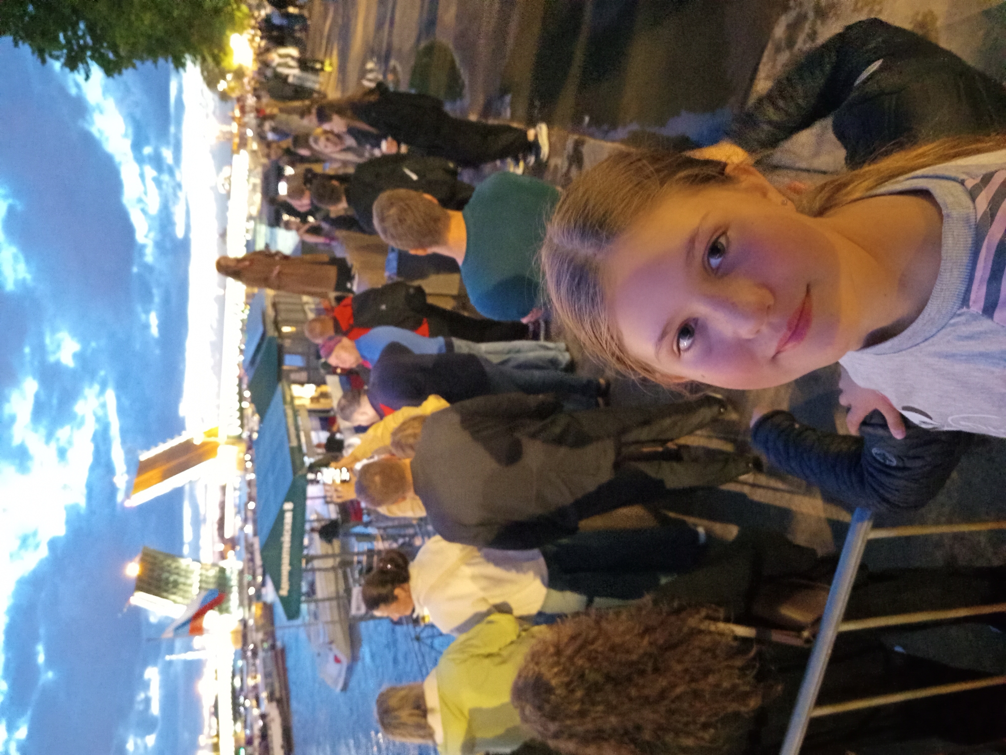 The image depicts a group of people gathered by a waterfront in Saint Petersburg, Russia, at dusk. In the background, two bridges are raised, revealing a Russian flag. A woman in a brown coat stands on a raised platform, observing the scene. The foreground is dominated by a young girl looking towards the camera, with her hand resting on a railing. Other individuals in the middle ground appear to be engaged in conversation and observing the raised bridges. The environment includes a body of water, a walkway, and trees. The sky is cloudy with hints of light from the setting sun.